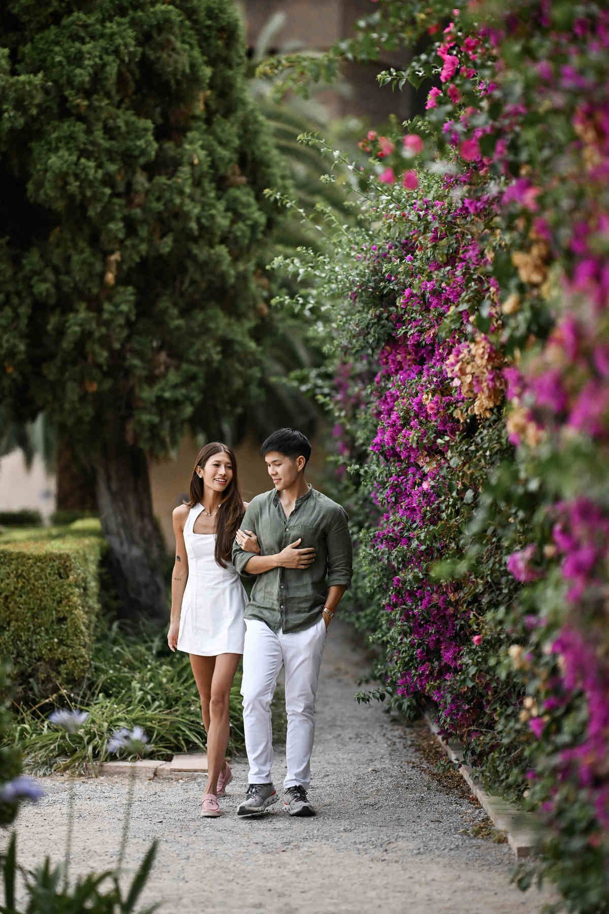 An international couple walking near bright pink flowers in Monforte Gardens after their secret proposal.