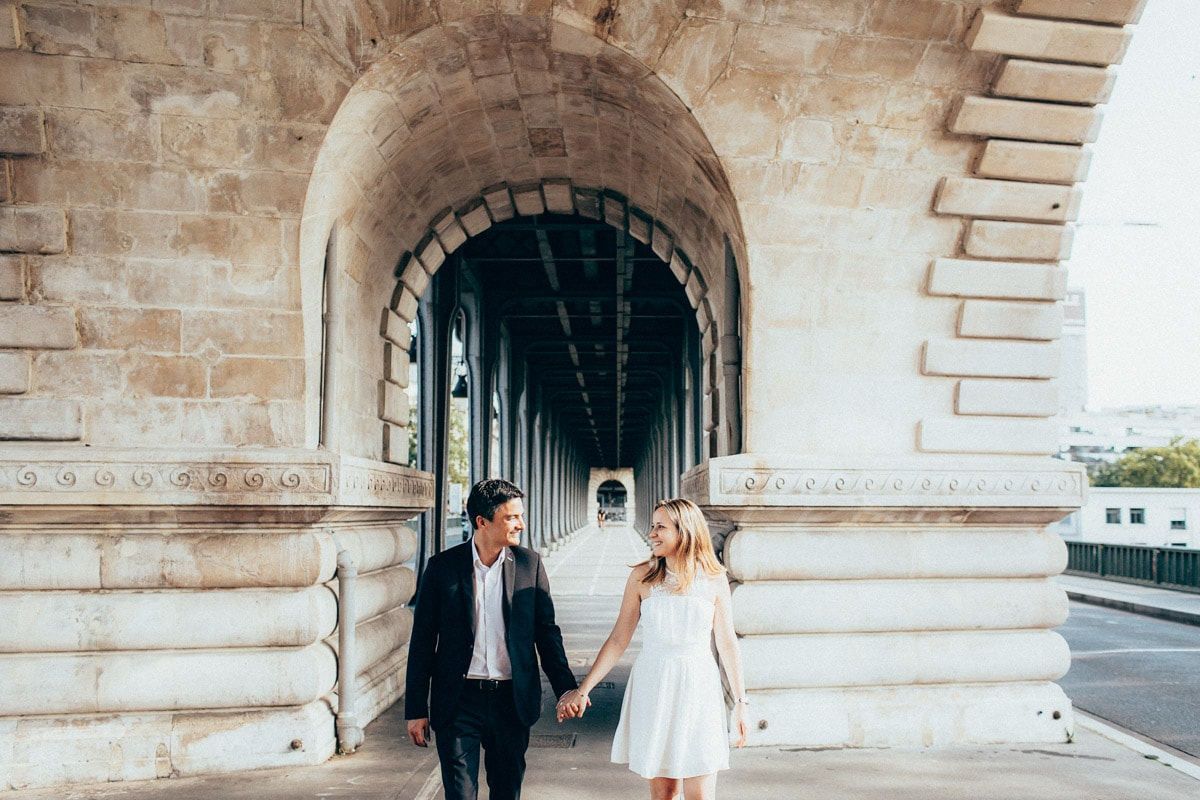 Happy engaged couple holding hands and walking under the historic stone arches of a Paris bridge during the daytime.