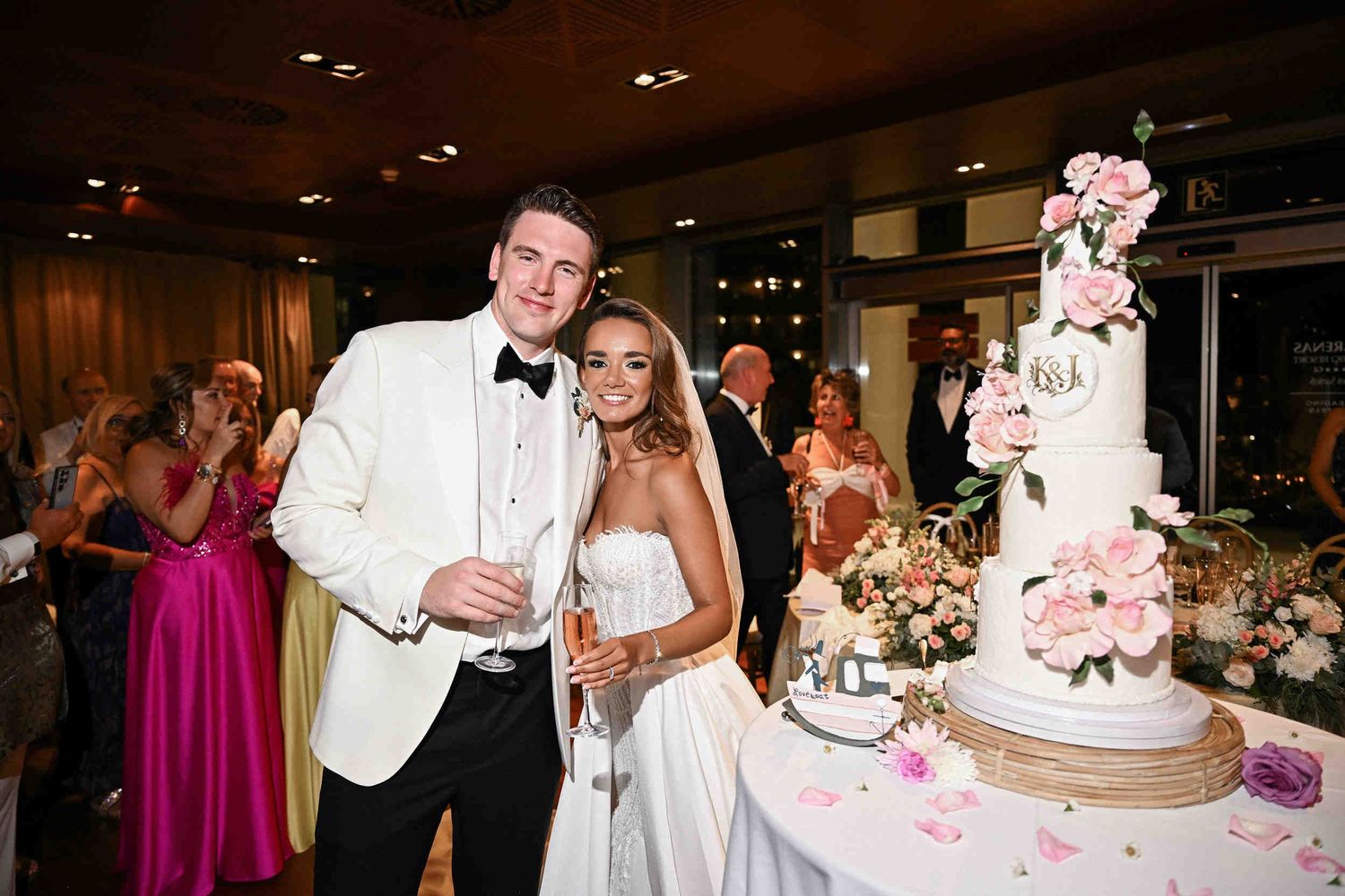 Newlyweds holding champagne glasses and smiling next to their floral tiered wedding cake at reception.
