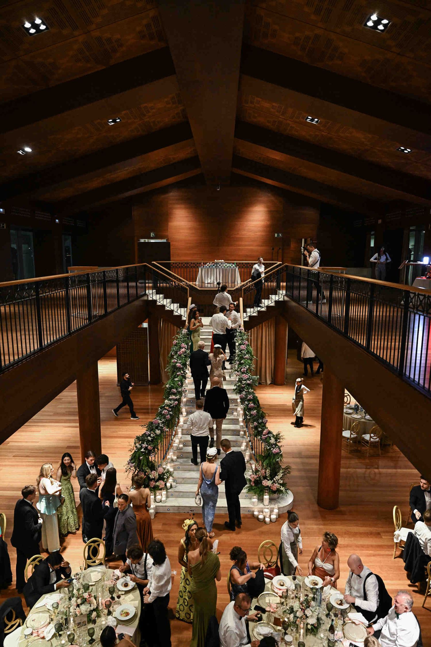 Wide view of a grand venue staircase decorated with pink florals and candles surrounded by banquet tables.