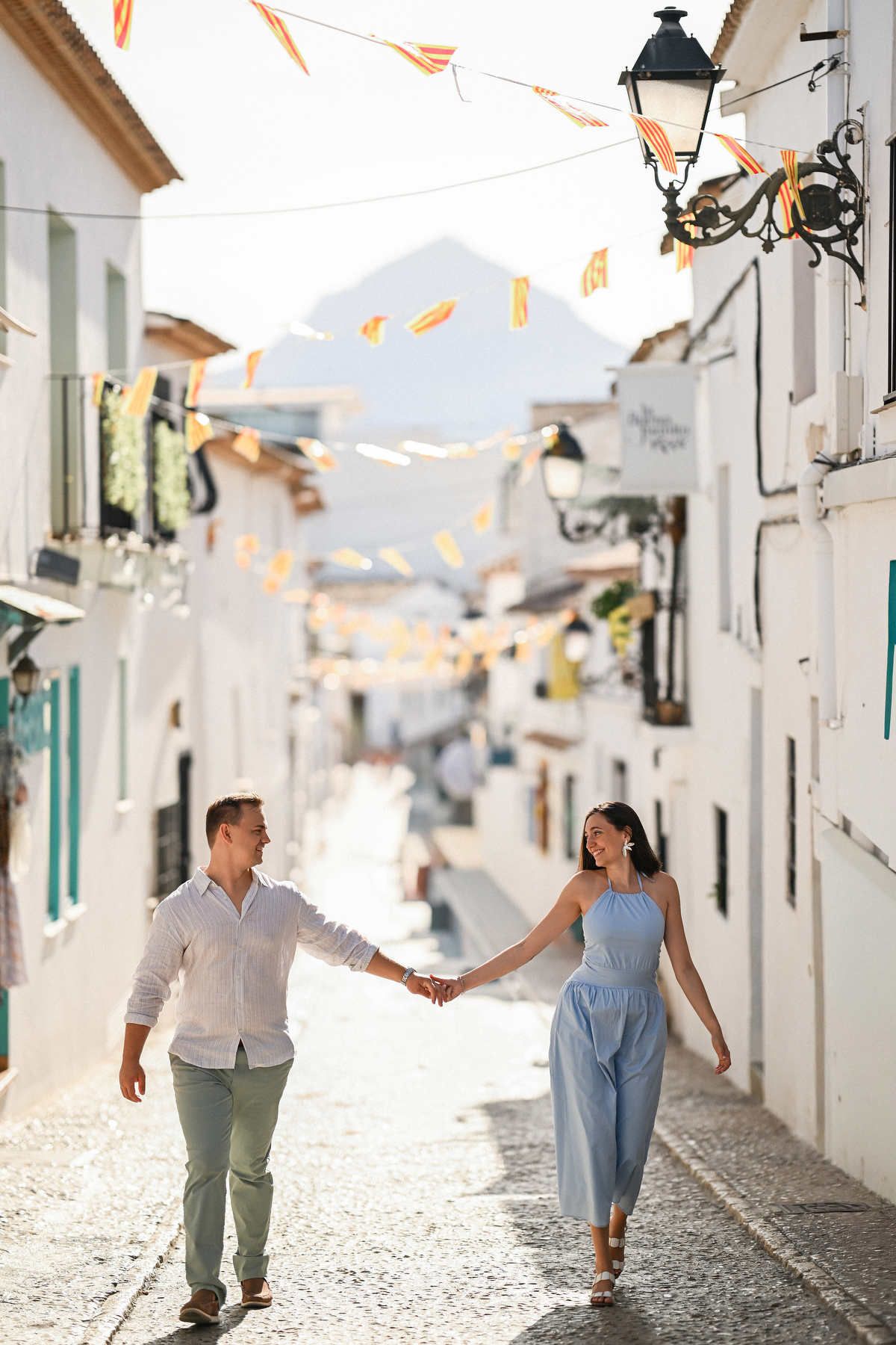 A happy international couple walks holding hands down a bright cobblestone street under festive bunting in Altea.