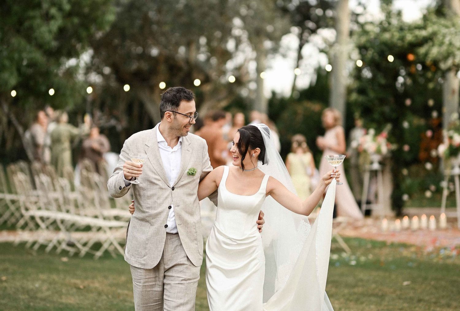 Happy newlyweds walking together with champagne glasses, smiling after their ceremony in the Mediterranean gardens
