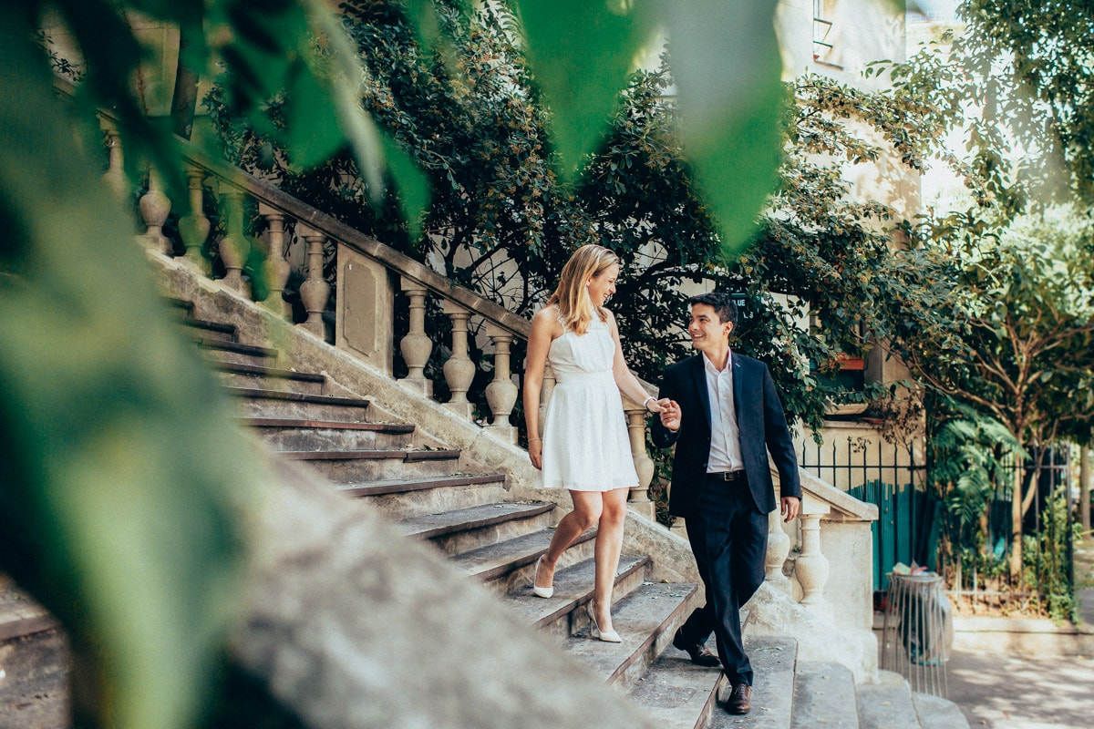 Engaged couple holding hands while walking down classic stone stairs framed by green leaves during a Paris pre-wedding session.