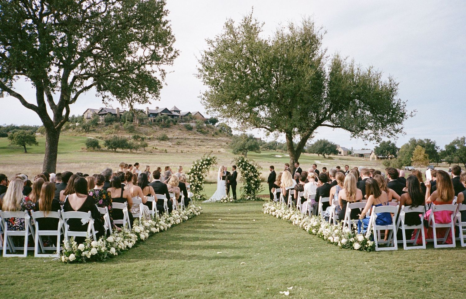Outdoor wedding ceremony site with white chairs lined up on a grassy hill under trees.