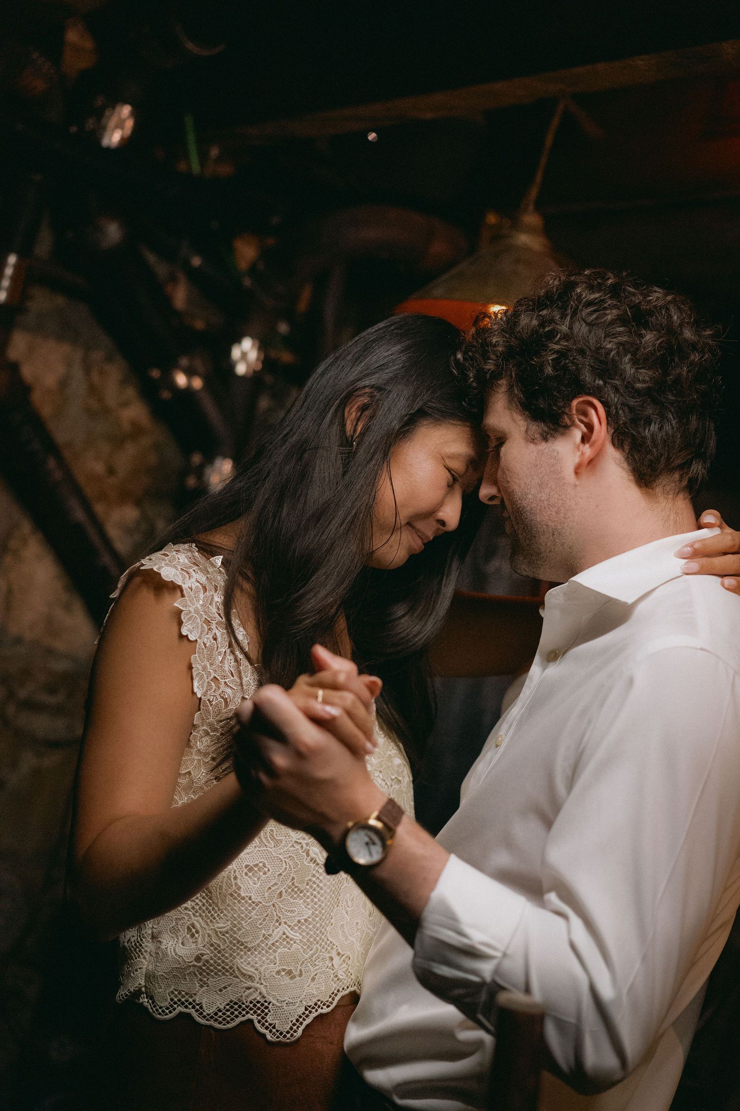 A couple shares an intimate slow dance, holding hands and touching foreheads in a warmly lit rustic venue.
