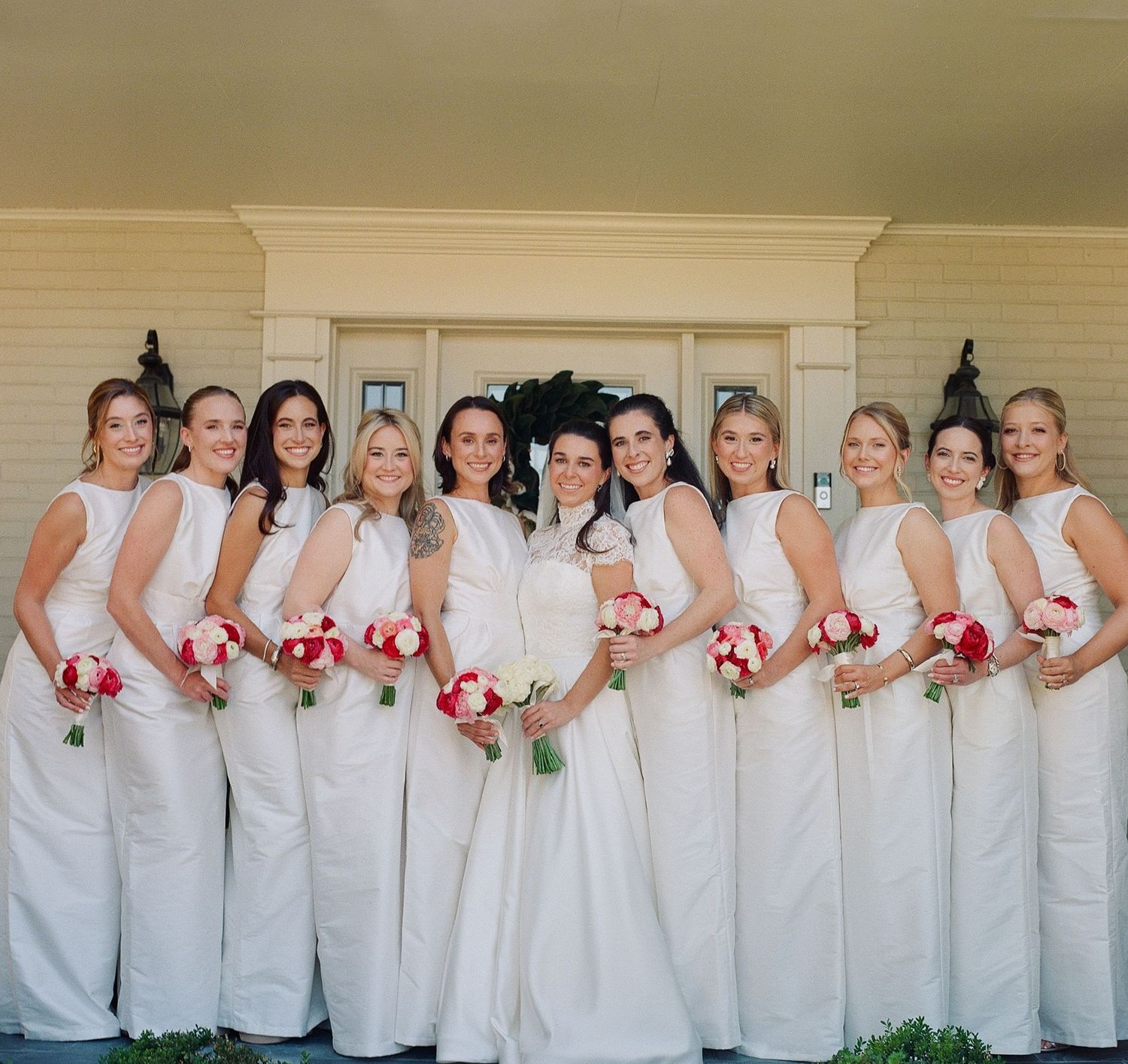 Wedding party members in white dresses holding red bouquets pose together against a light wall.