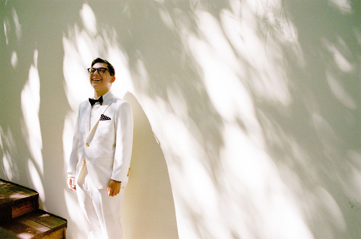 A figure in white formal attire stands against a light wall with dramatic shadow patterns.