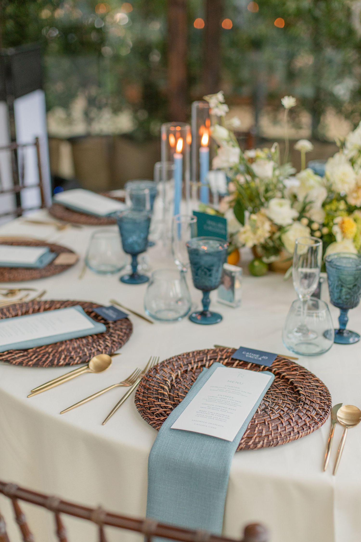 Table setting with blue glasses, gold cutlery, and woven placemats on white tablecloth.
