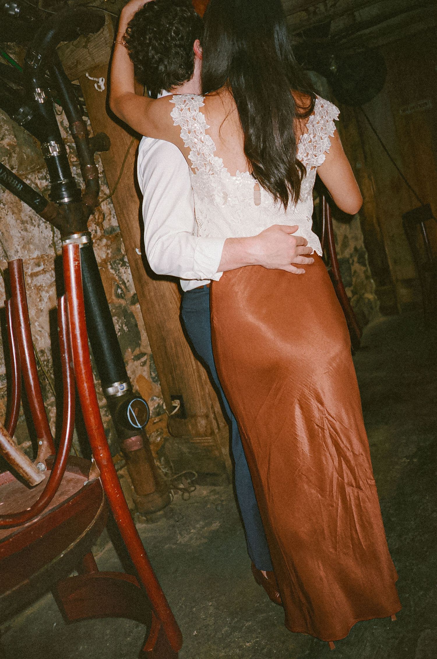 Couple dancing closely in a dimly lit rustic venue, woman in lace top and rust satin skirt, man in white shirt.