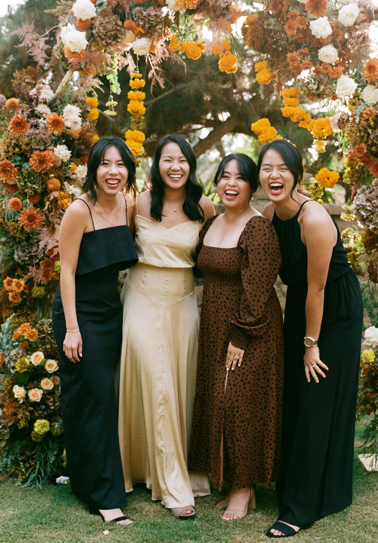 Four women smiling in front of a floral arch with orange and yellow blooms at an outdoor wedding celebration.