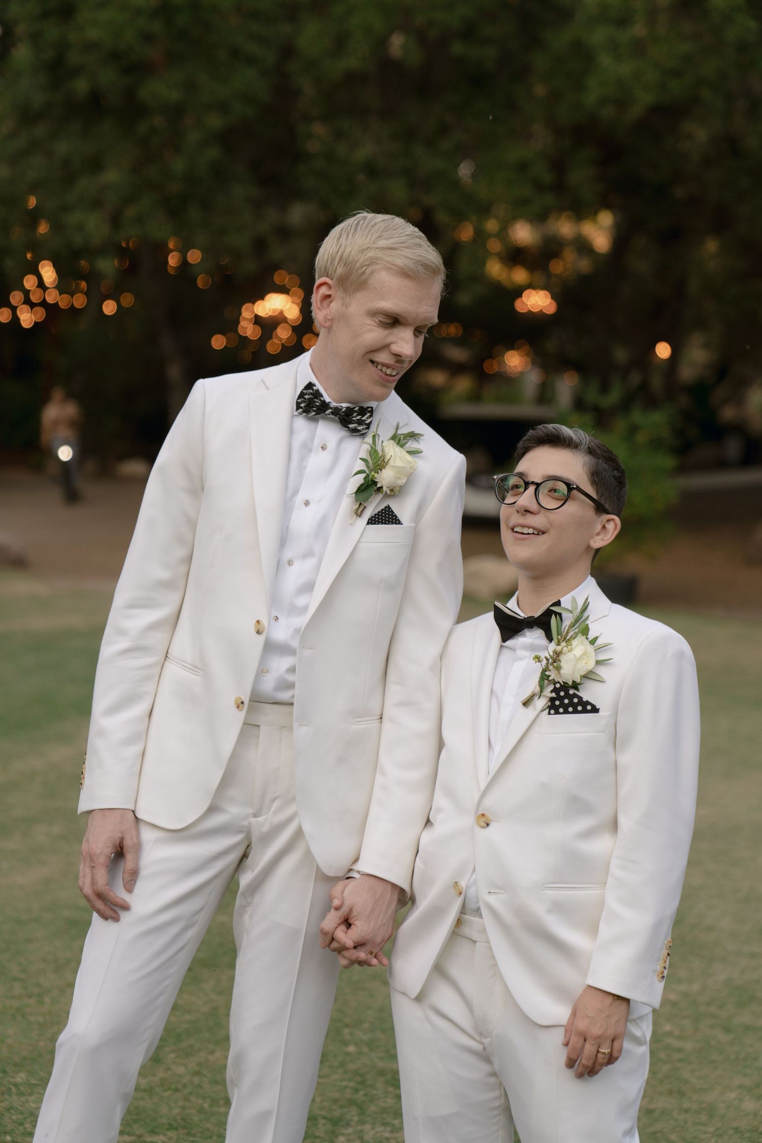 A couple in matching white tuxedos share a romantic moment outdoors.