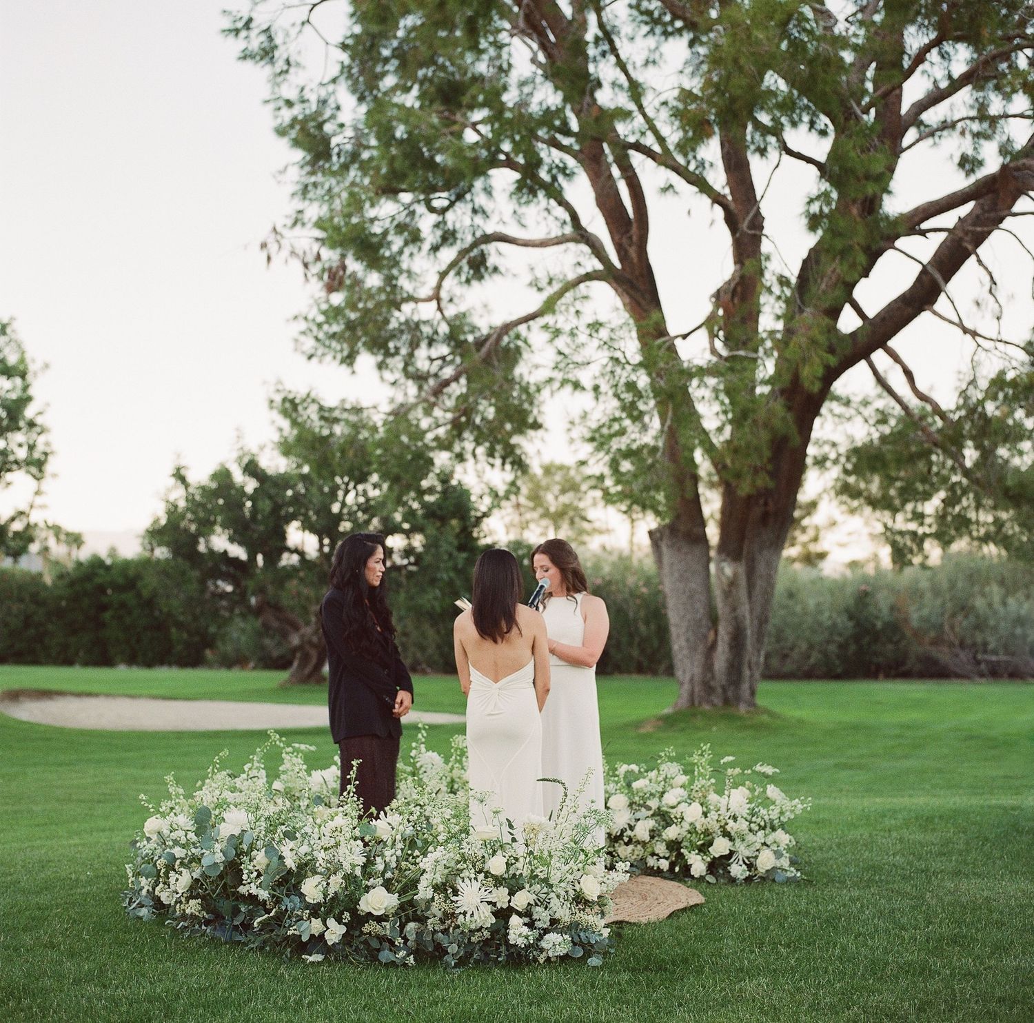 Wedding ceremony participants walking on a grass lawn surrounded by white flowers.