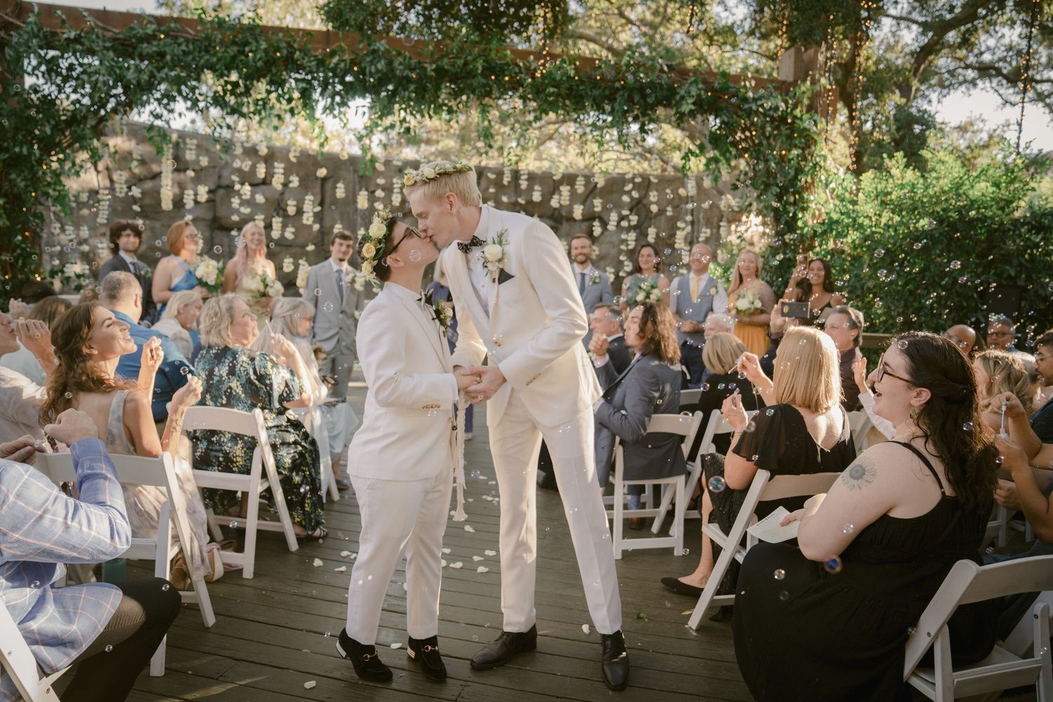 Outdoor wedding ceremony guests seated in white chairs under a canopy of trees.