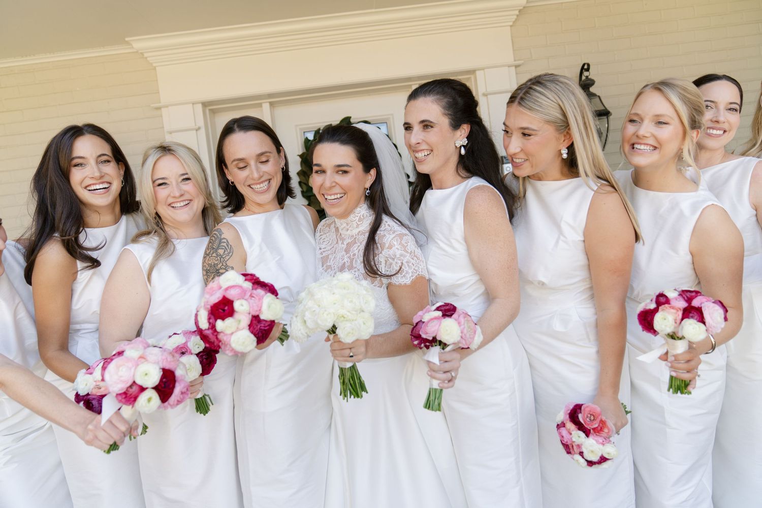 Bridesmaids in elegant white dresses pose together holding pink and white bouquets.