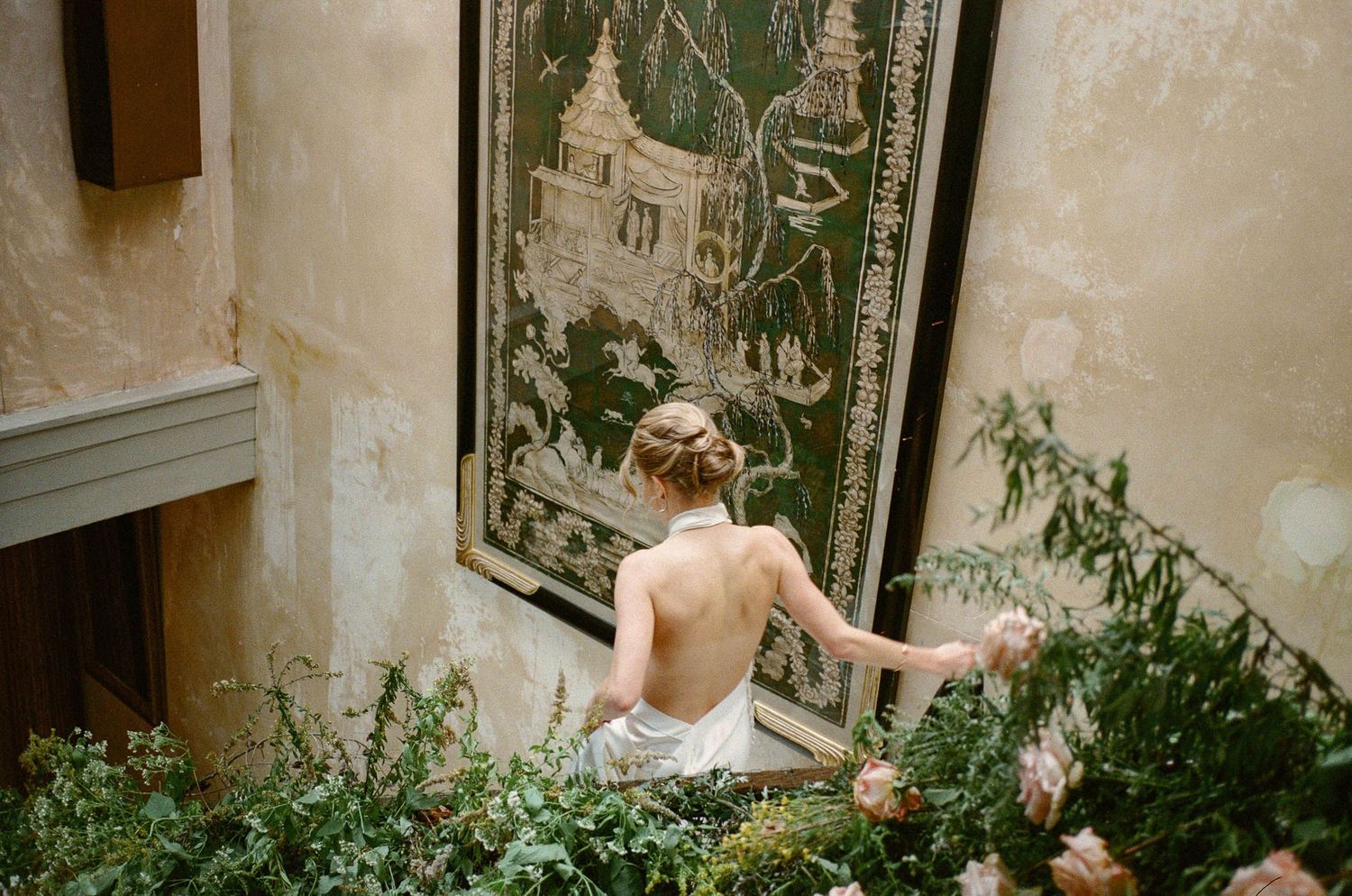 A figure in white sits near a decorative wall panel surrounded by lush greenery in an elegant interior setting.