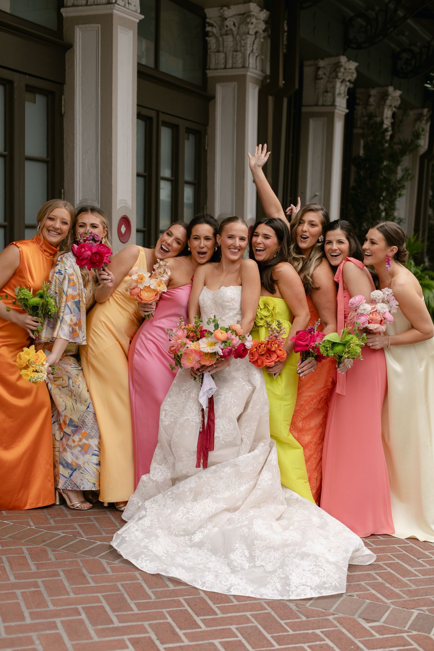 Bridesmaids in colorful orange and pink dresses pose joyfully with bride in white gown on brick patio.