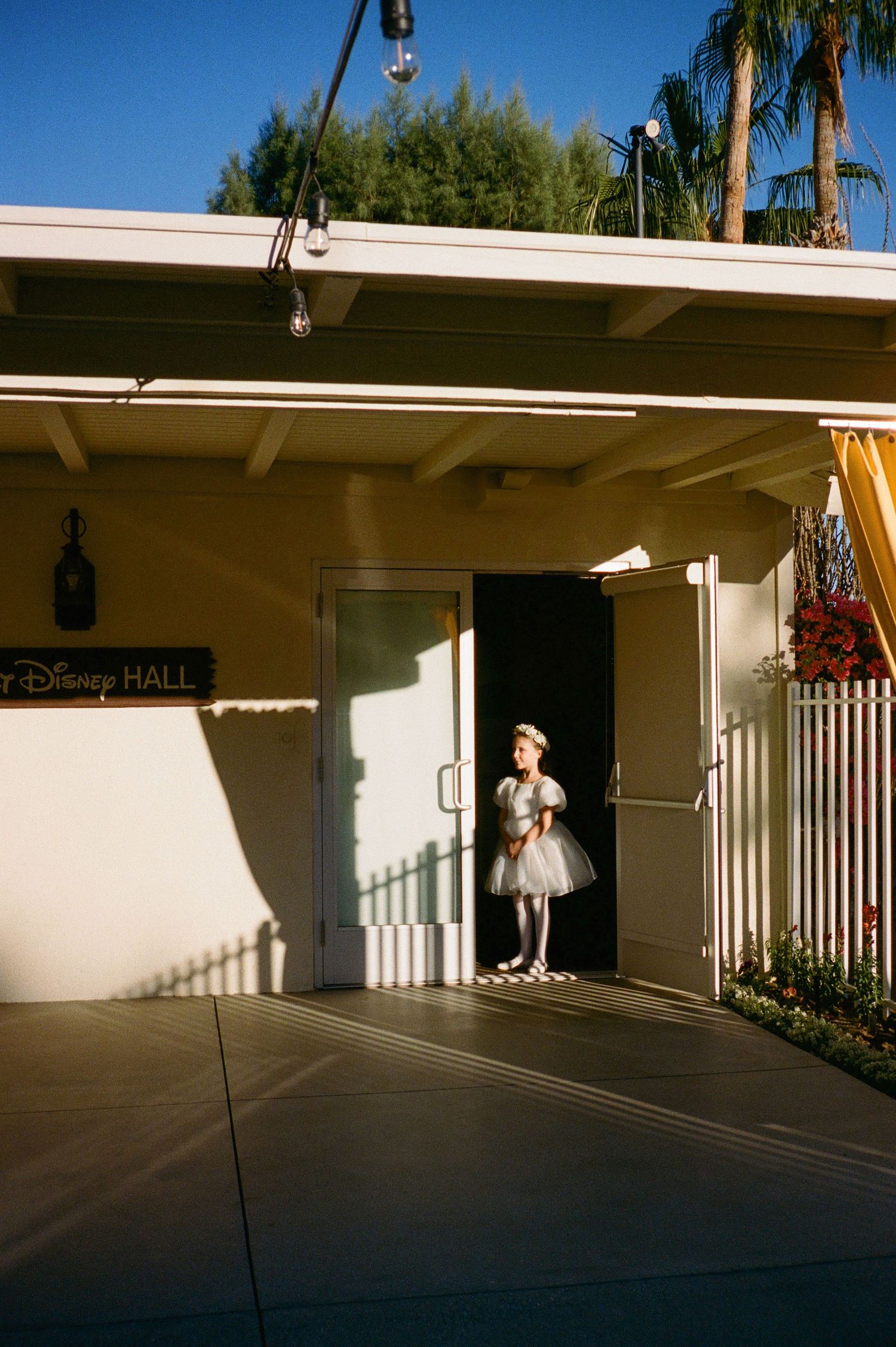 A young girl in a white tutu dress stands in a doorway at Disney Hall, bathed in warm sunlight.