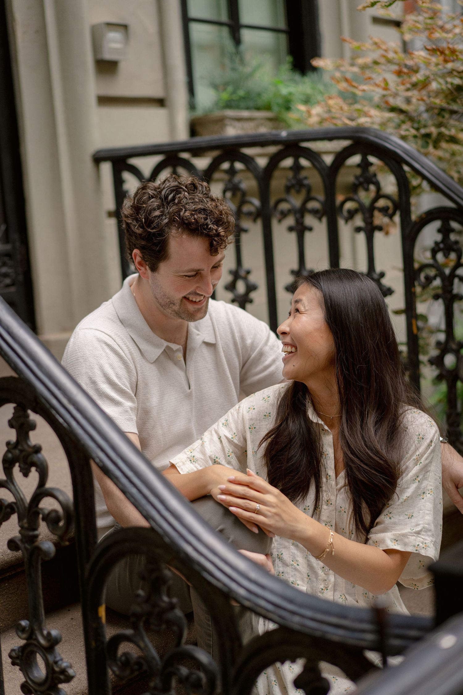 A smiling couple shares a joyful moment on ornate iron steps outside a classic building.