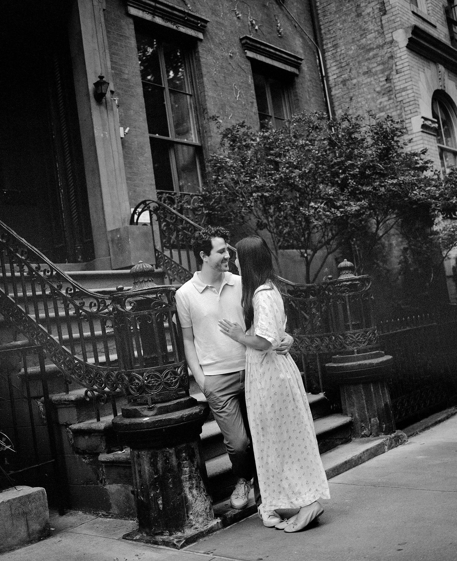 Black and white photo of a couple embracing and smiling outside a classic brownstone building with ornate iron railings.