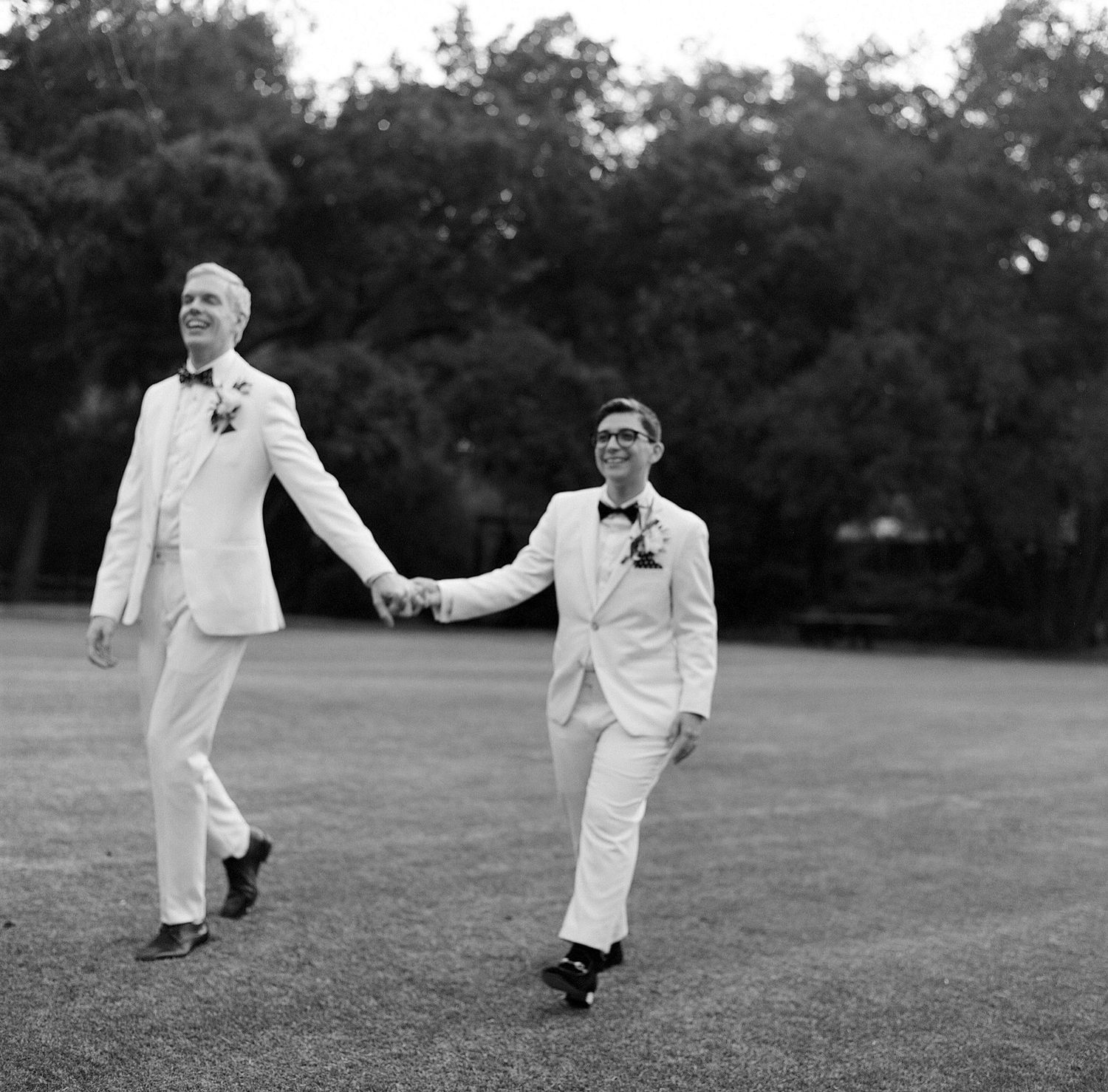 Two people in white formal attire playfully pose on a grassy lawn at sunset.