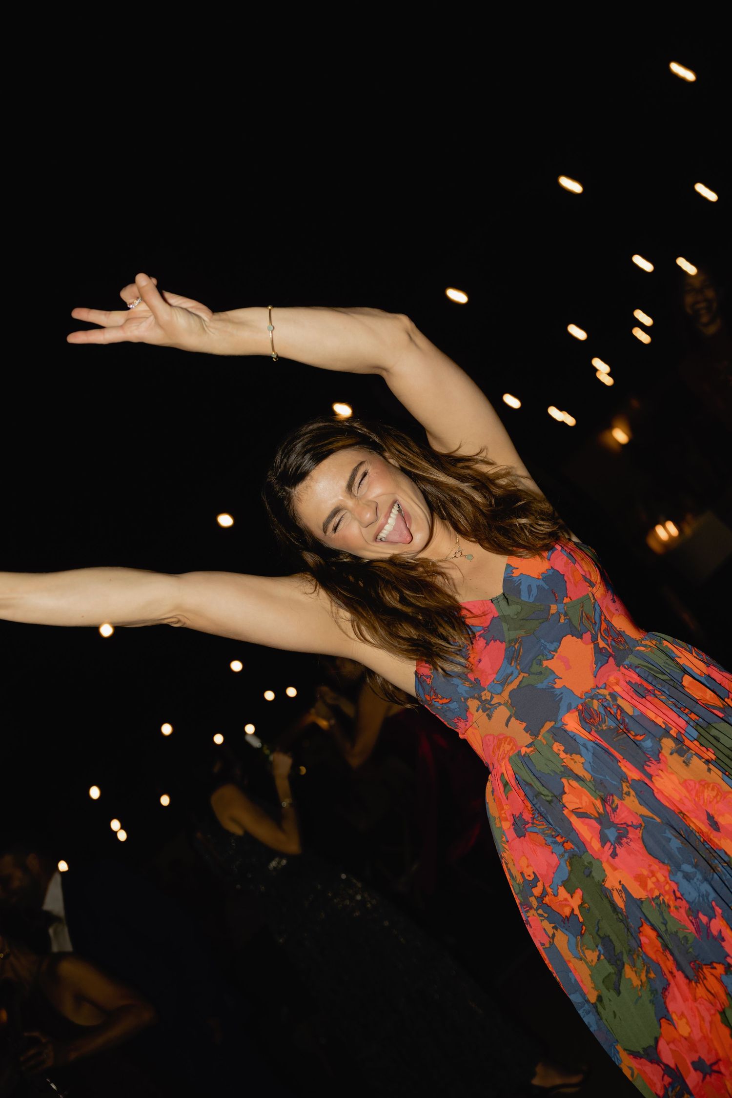 Person in a floral dress twirling with arms outstretched under twinkling lights at night.