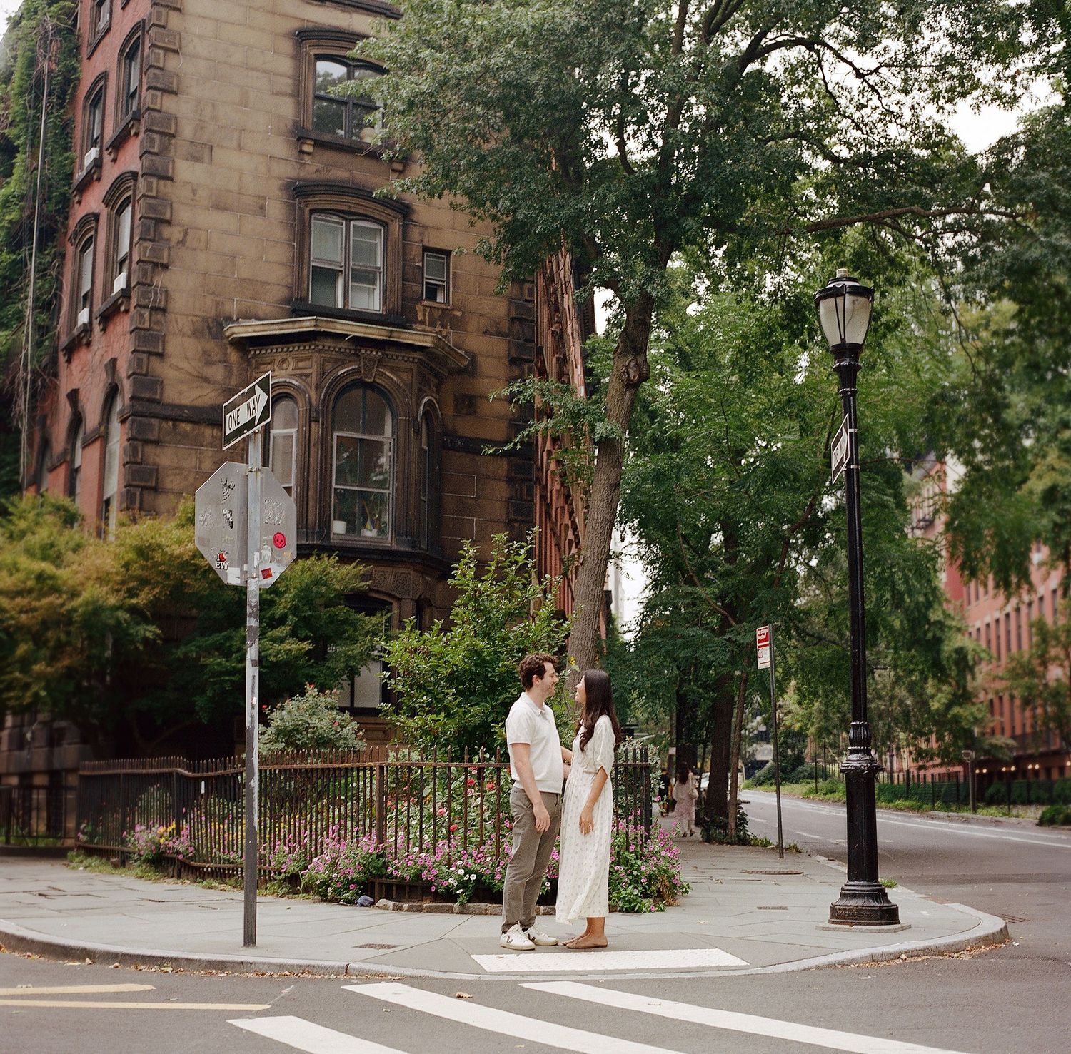 Couple standing at a tree-lined NYC street corner near historic brownstone buildings on a summer day.