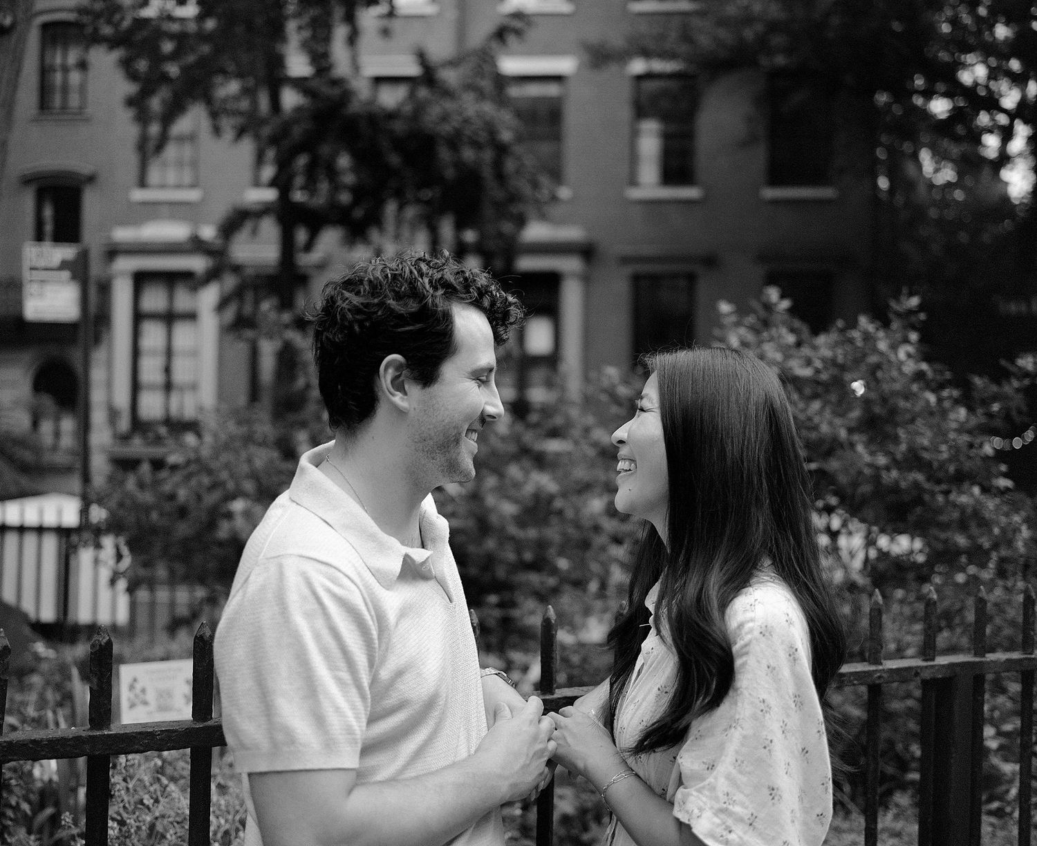 A smiling couple gazes at each other outdoors in a black and white urban photo with brownstones in the background.