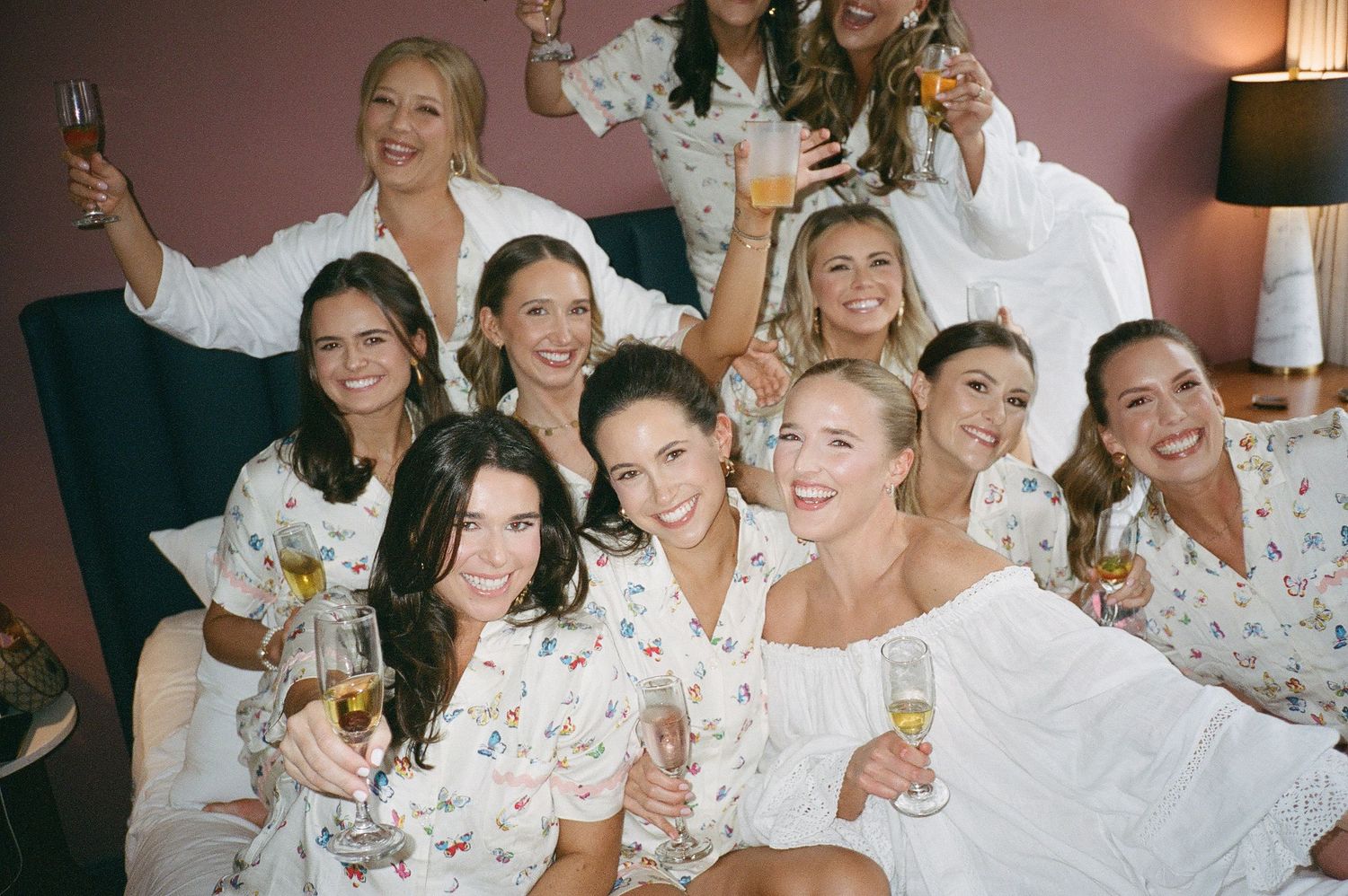 Wedding party in floral robes celebrates with drinks and laughter in a hotel room.