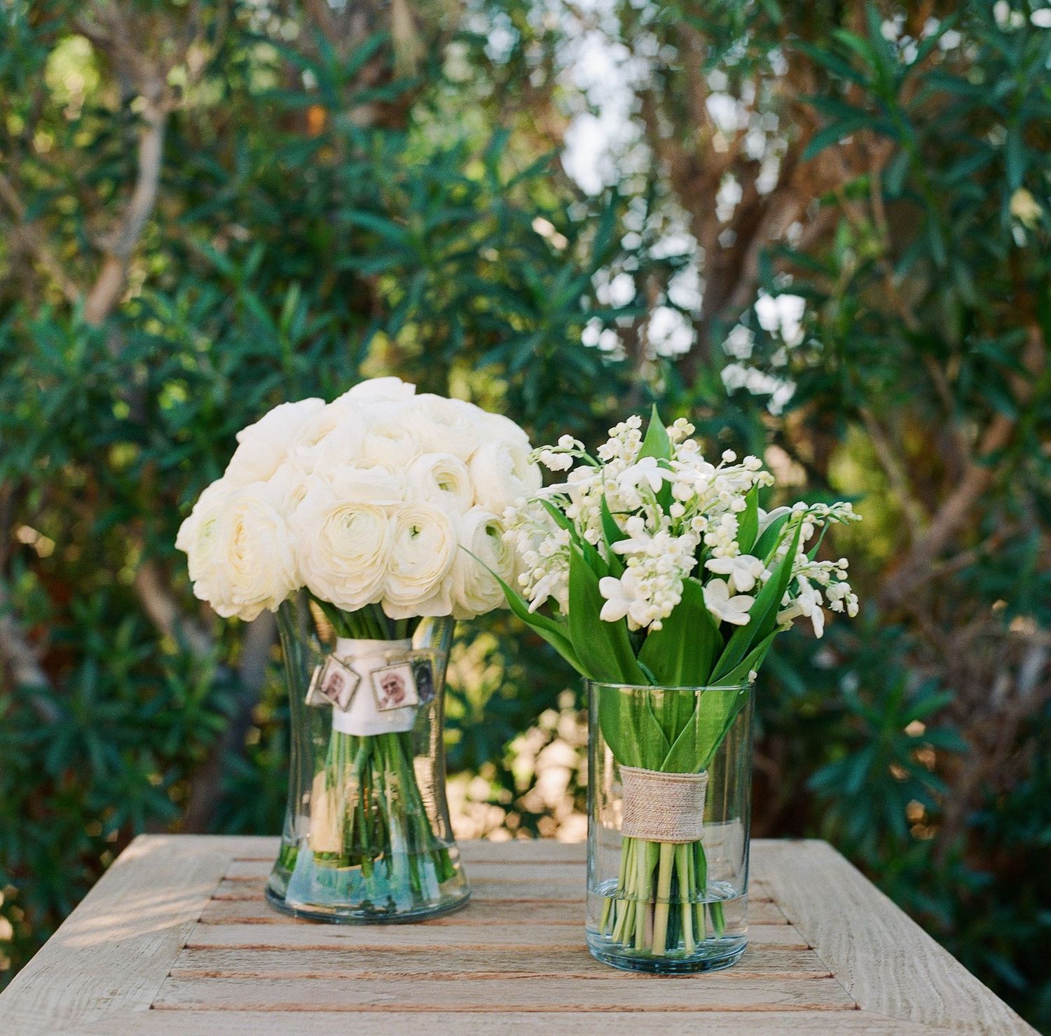 White roses and flowers arranged in glass vases on a rustic wooden table outdoors.
