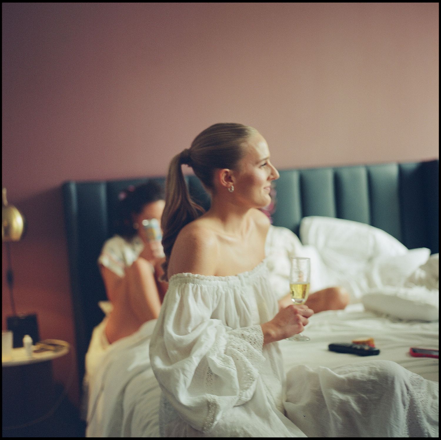 Woman in white off-shoulder dress holding champagne glass, sitting on bed in pink room.