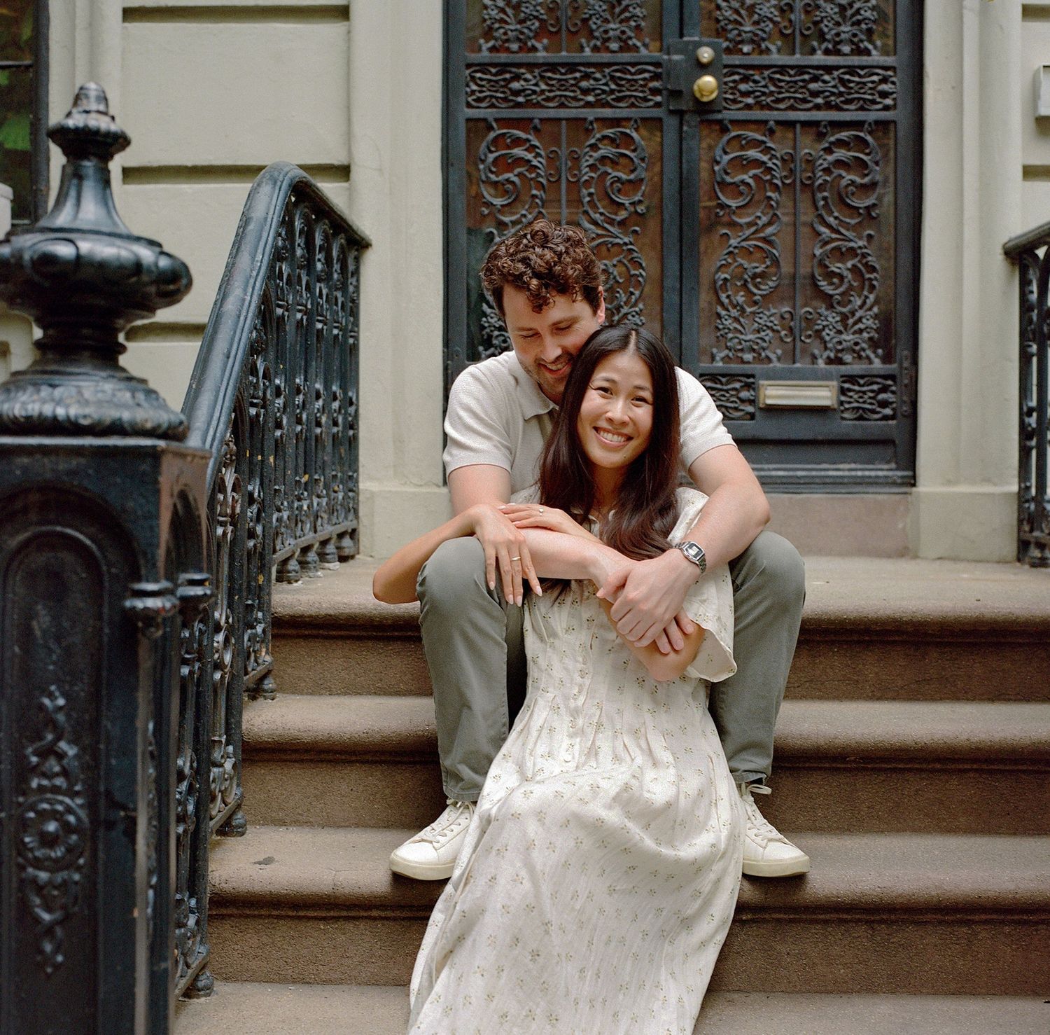 Couple sitting on brownstone steps embracing, woman in white dress, man in casual outfit, urban NYC backdrop.