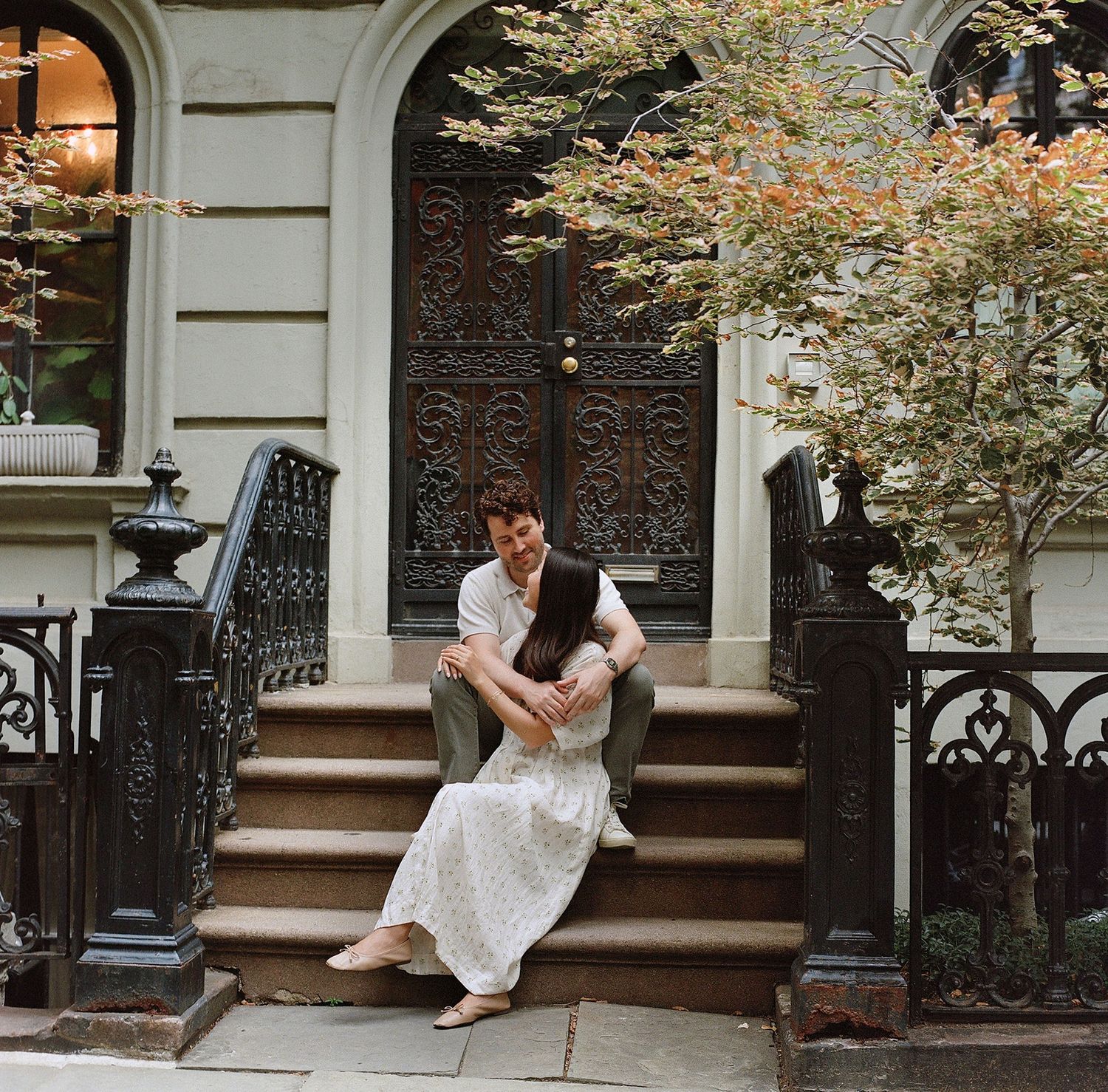 A couple sits embracing on brownstone steps beside ornate iron railings and an autumn tree in an urban setting.