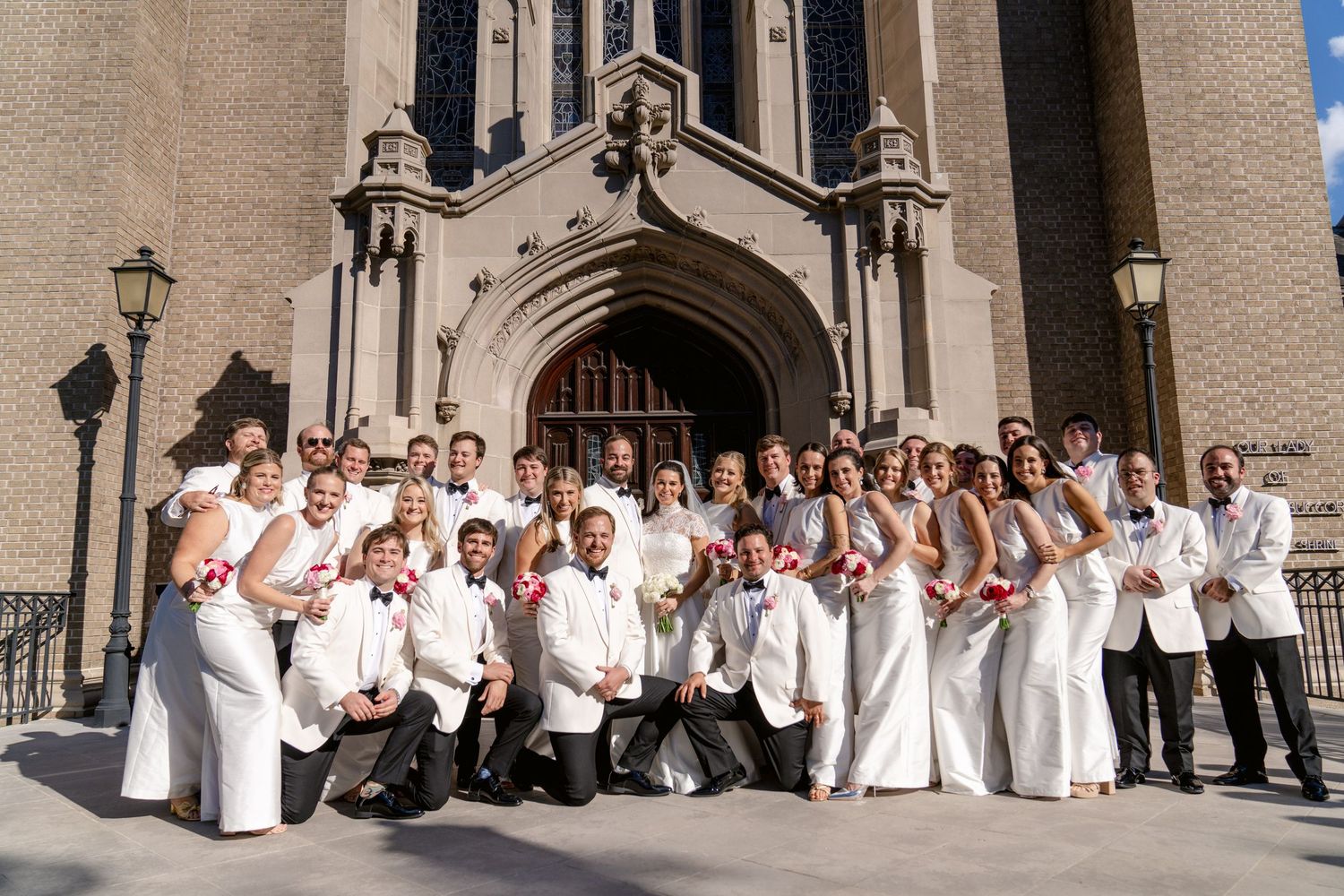 Large wedding party in white attire poses for group photo in front of stone church building.