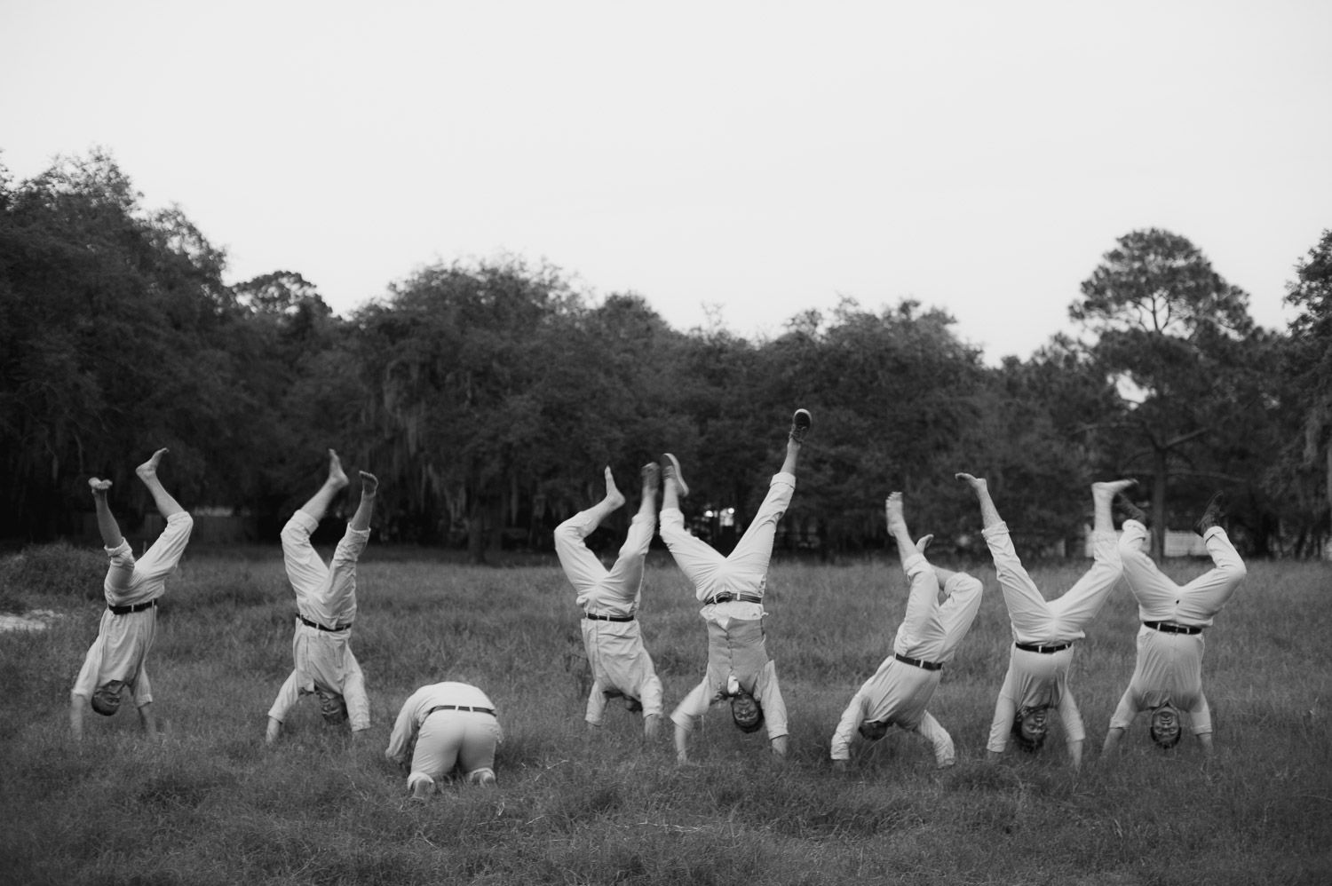 Wedding party members lying on grass doing synchronized poses in black and white photography.