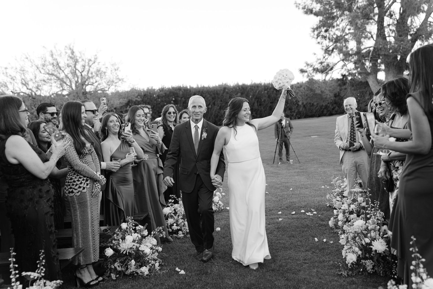 Black and white photo of wedding ceremony with guests applauding along flower-lined aisle.