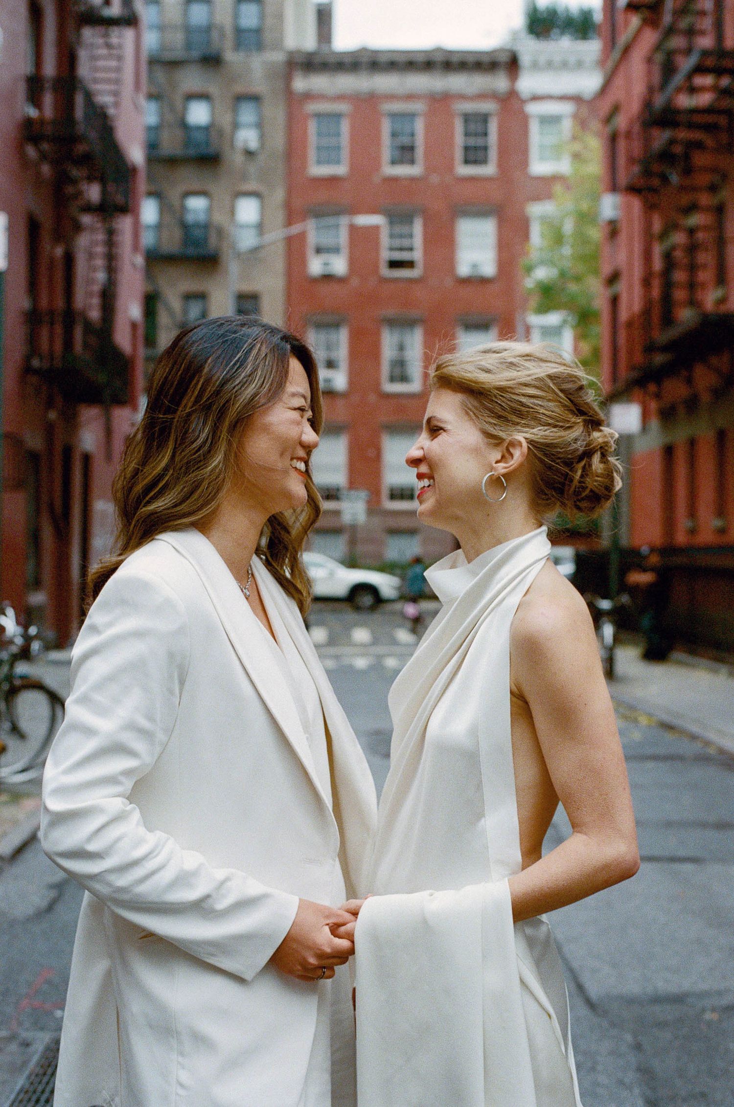 Two people in white outfits share joyful moments on a city street with brownstone buildings.