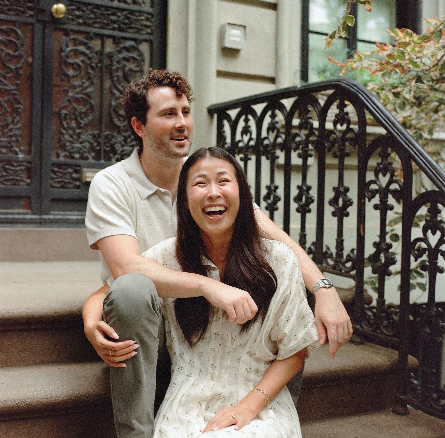A laughing couple sits on brownstone steps in an urban setting, the woman in a floral dress and man in a polo shirt.