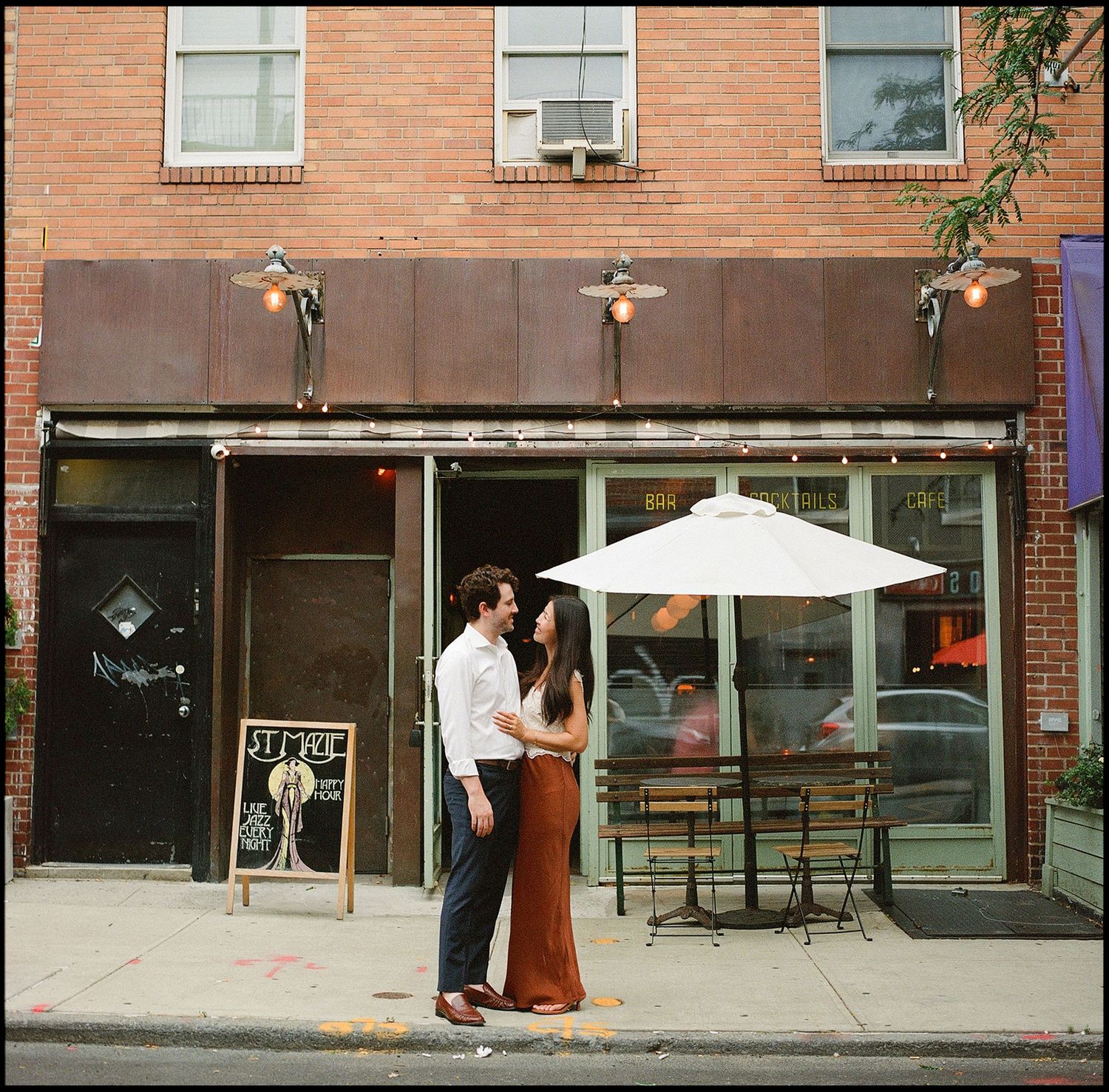 Couple sharing a romantic moment outside a brick storefront cafe with a white patio umbrella on a city sidewalk.