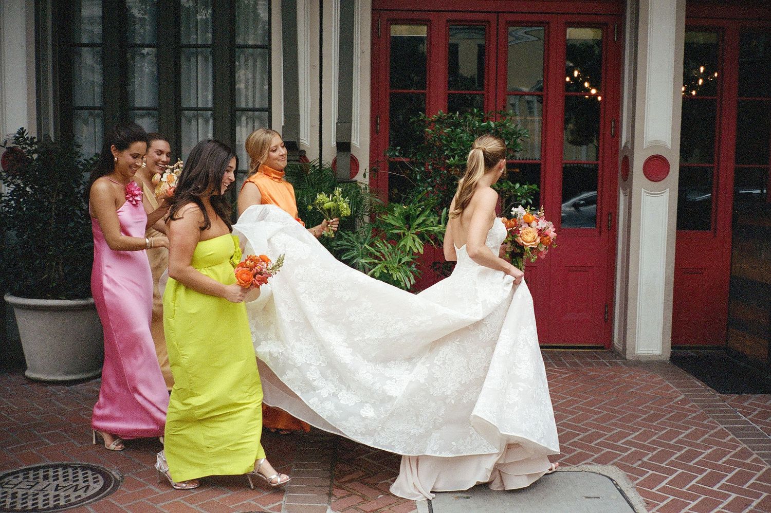 Wedding party helps bride with dress on brick sidewalk outside building with red doors.
