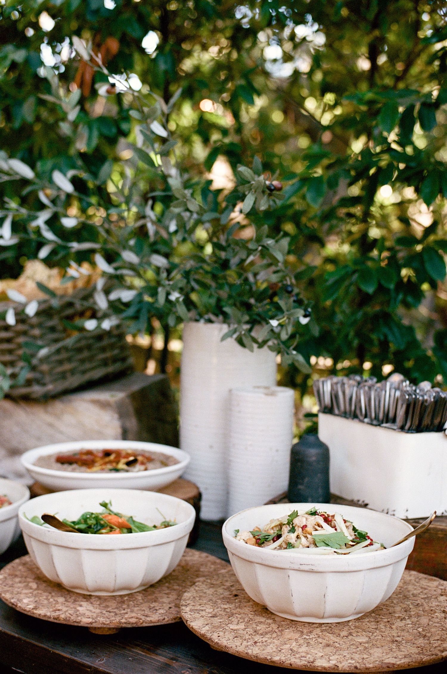 White serving bowls filled with salads on a rustic table with greenery decor.