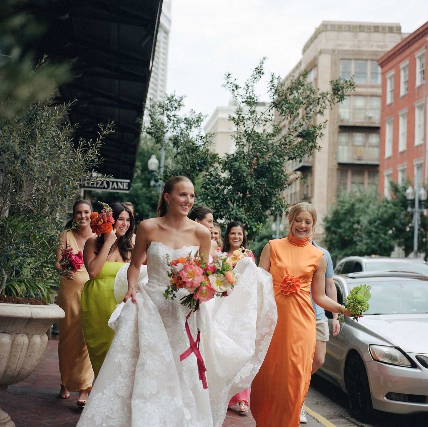 Wedding party in orange and yellow dresses walks along urban sidewalk during photo session.