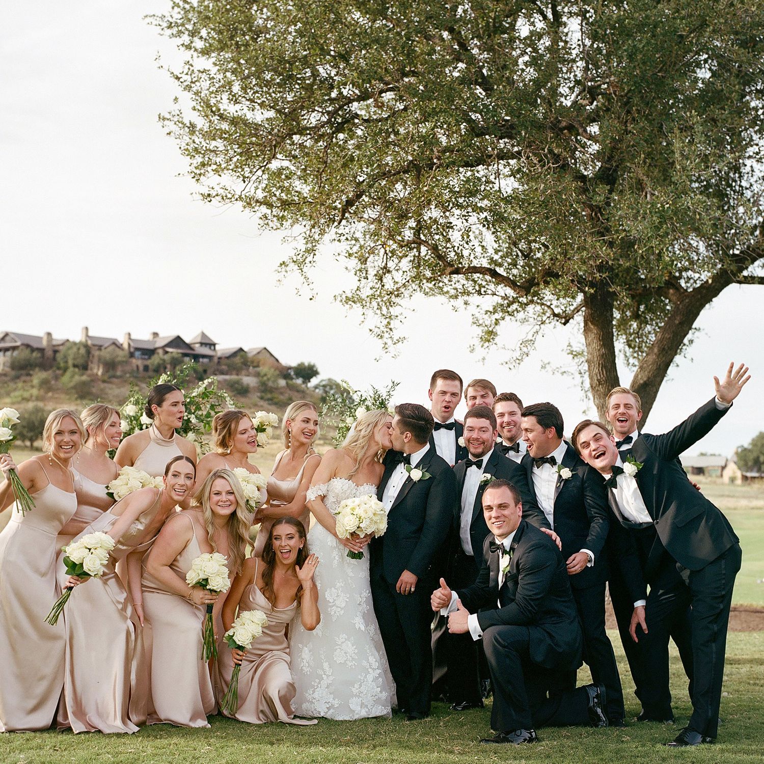 Wedding party poses creatively outdoors with groomsmen lying on grass and bridesmaids standing in blush gowns.