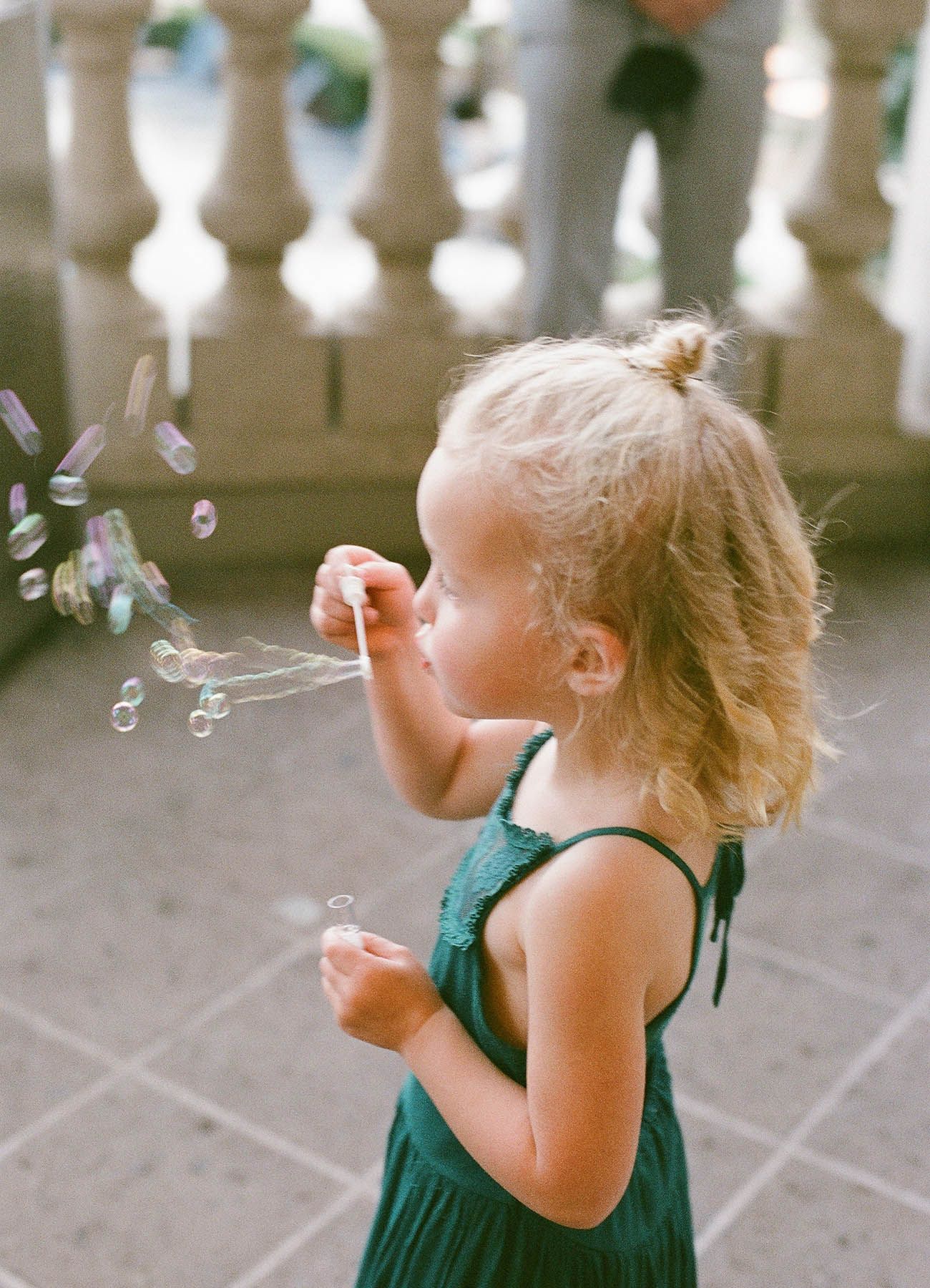 A young blonde girl in a green dress blows bubbles outdoors, captured in a candid film photography moment.
