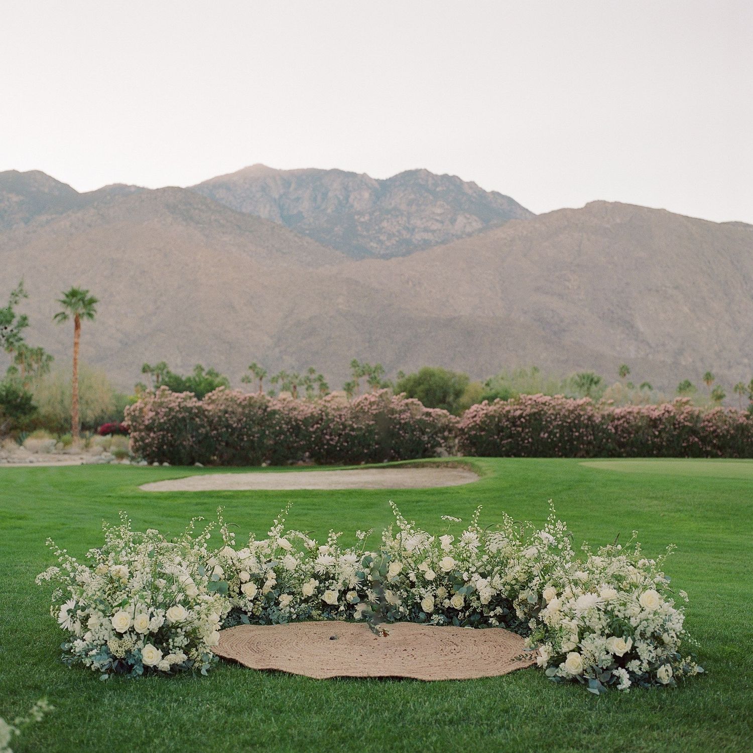 White floral ceremony arch arrangement on a green lawn with desert mountains in the background.