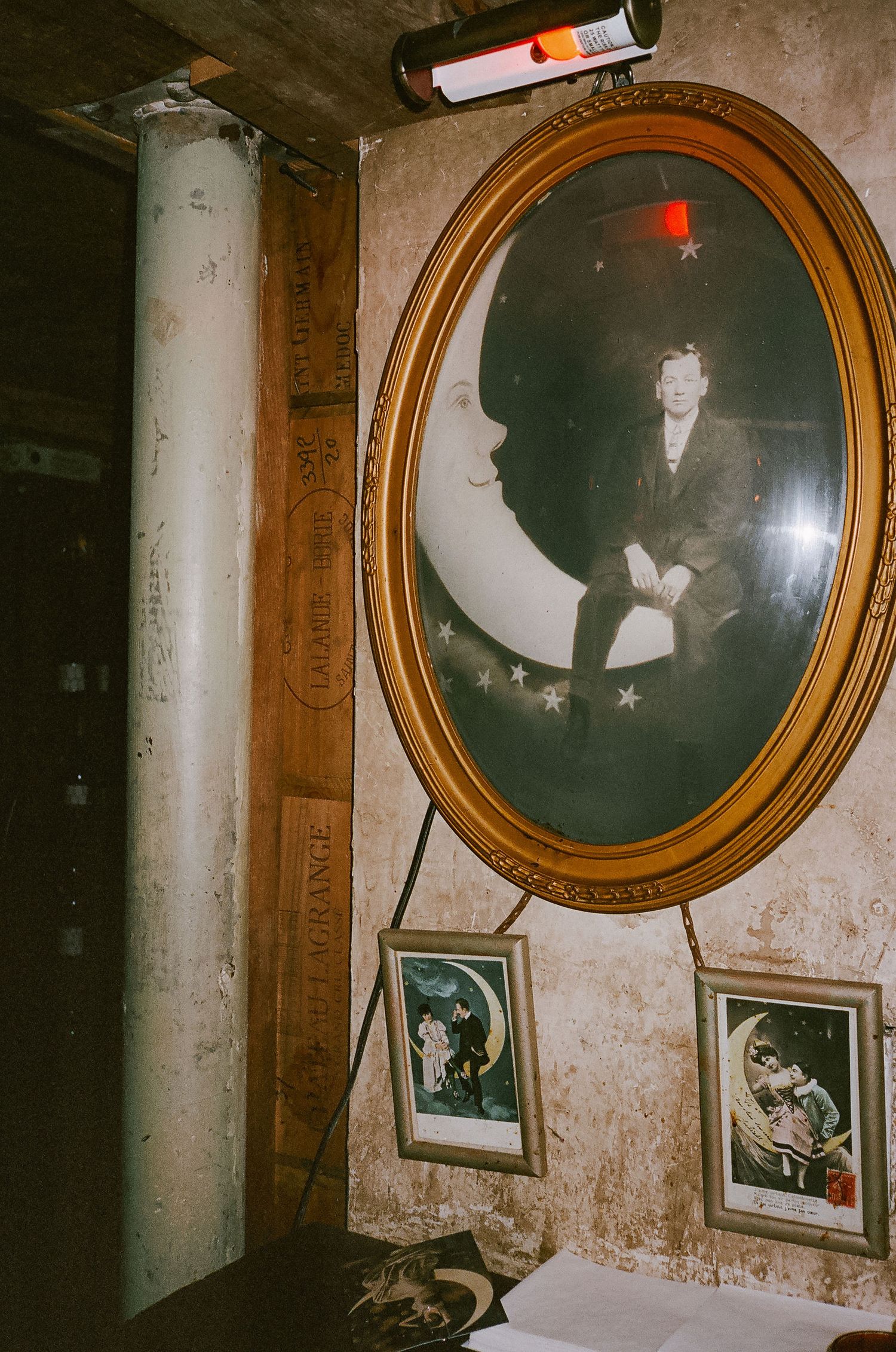 Vintage oval gold-framed portrait of a man sitting on a crescent moon, displayed on a distressed wall with smaller framed prints.