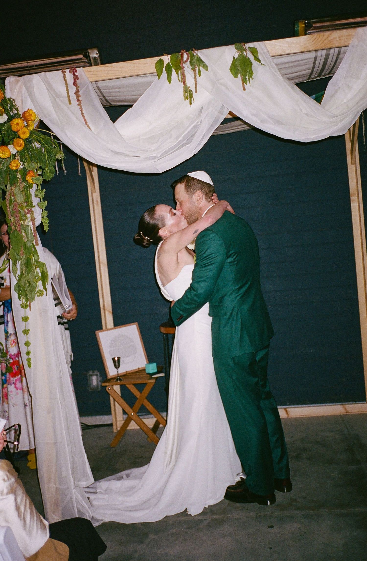 Bride and groom share first kiss under floral chuppah, groom in green suit wearing kippah, bride in white gown.