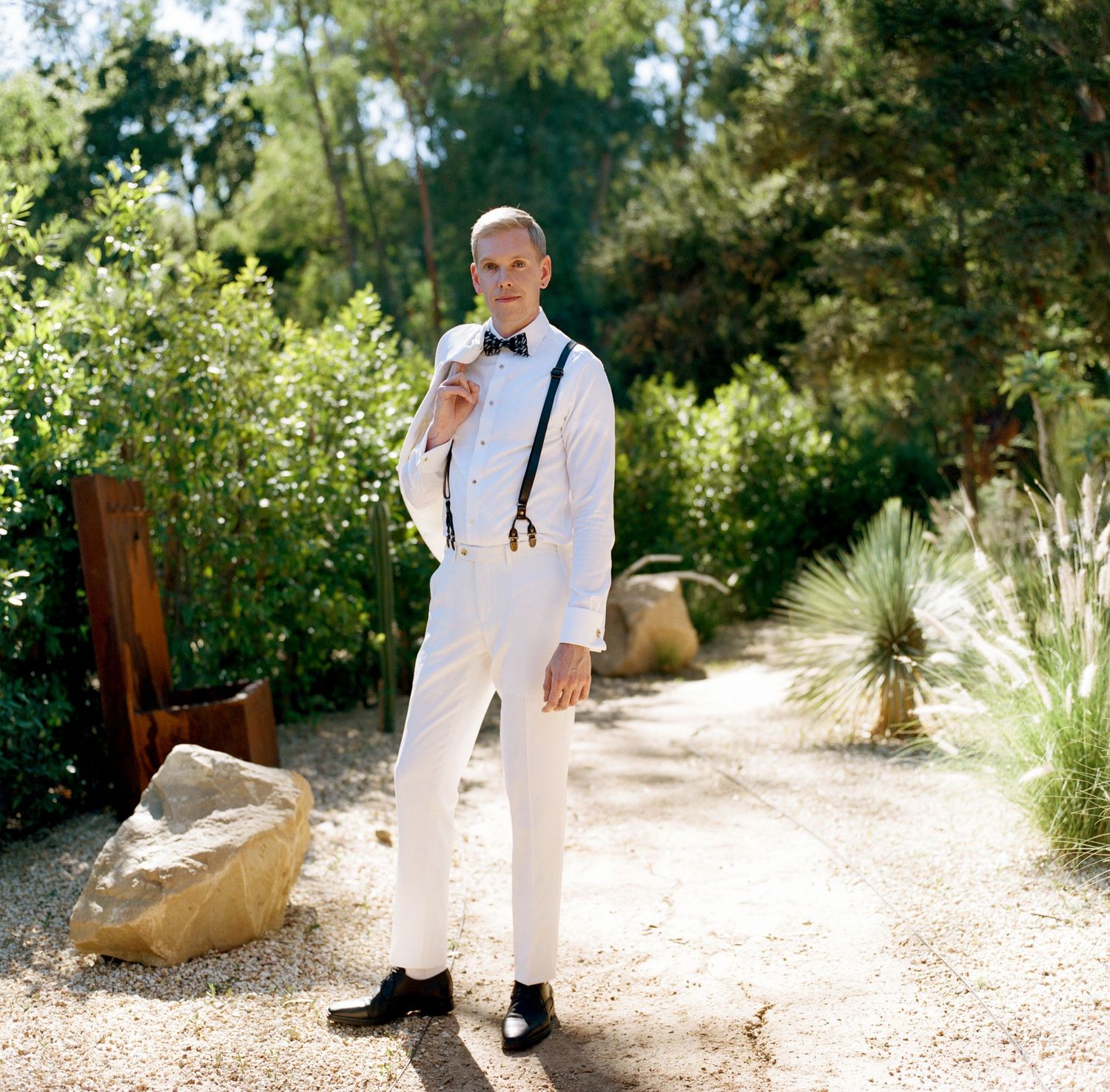 Someone in an all-white formal outfit walks along a garden path with natural sunlight filtering through trees.