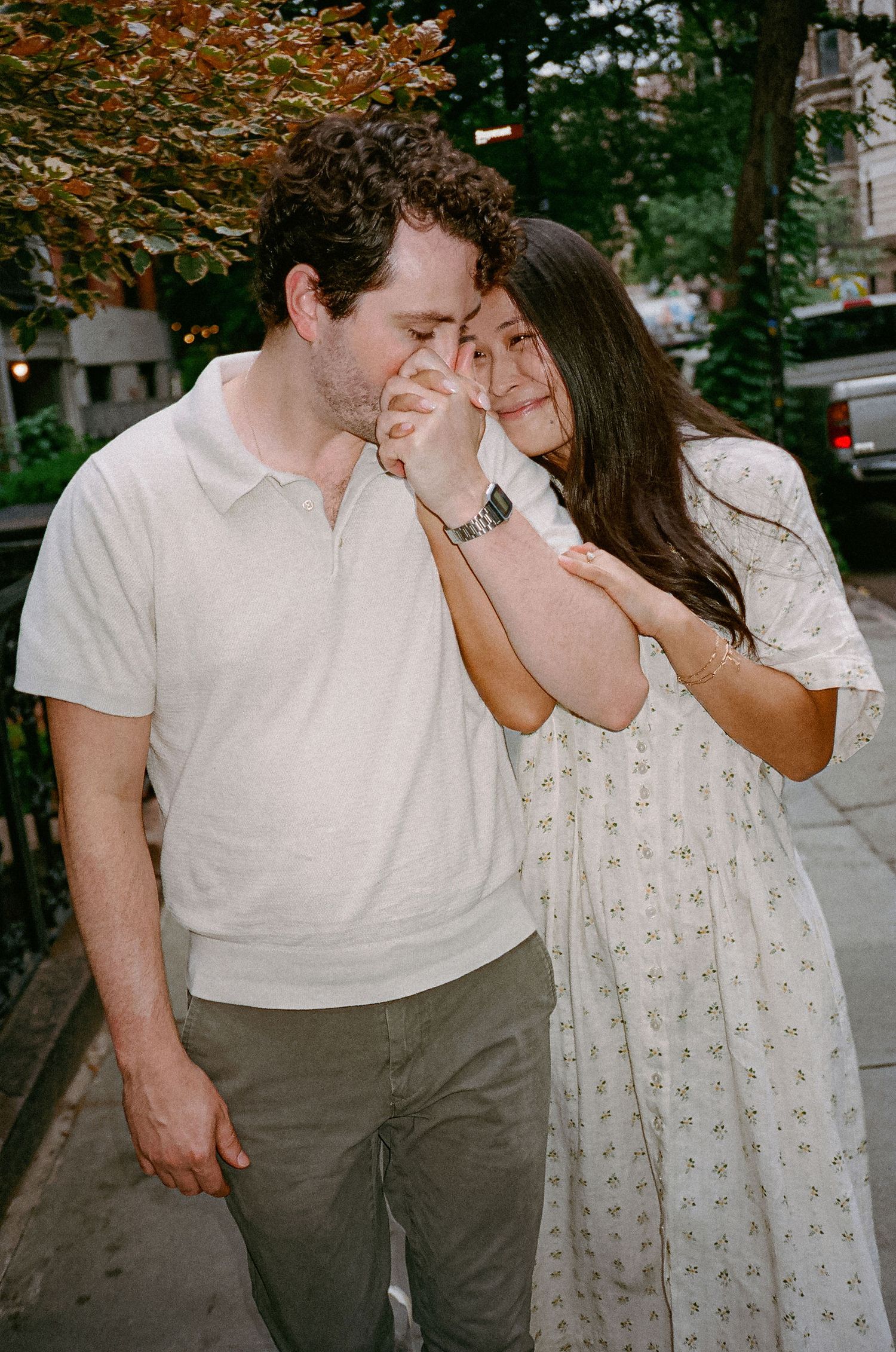 A couple shares a tender moment on a city sidewalk, the woman leaning into the man while he kisses her hand.