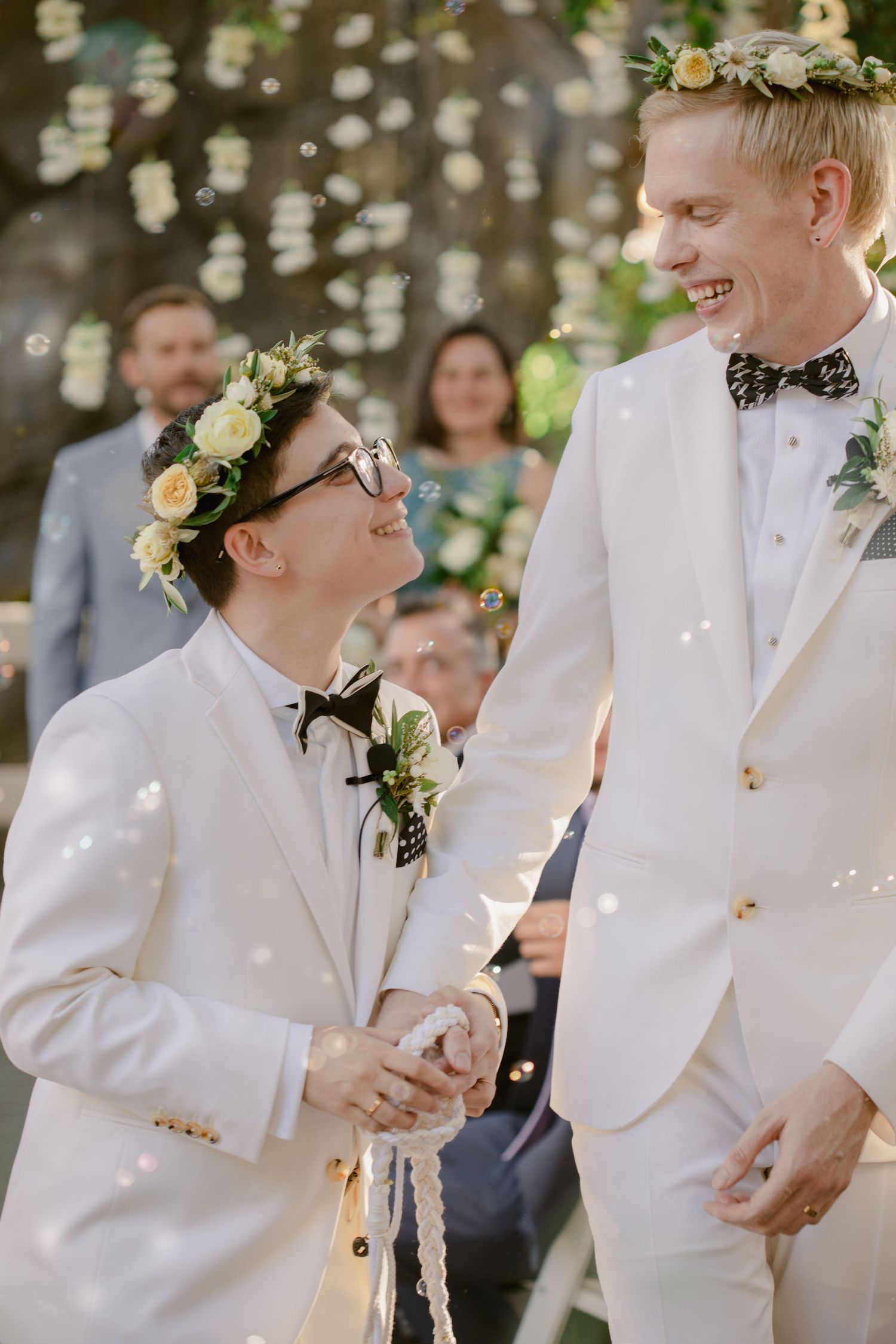 Wedding ceremony participants in white suits and bow ties share a joyful moment outdoors.