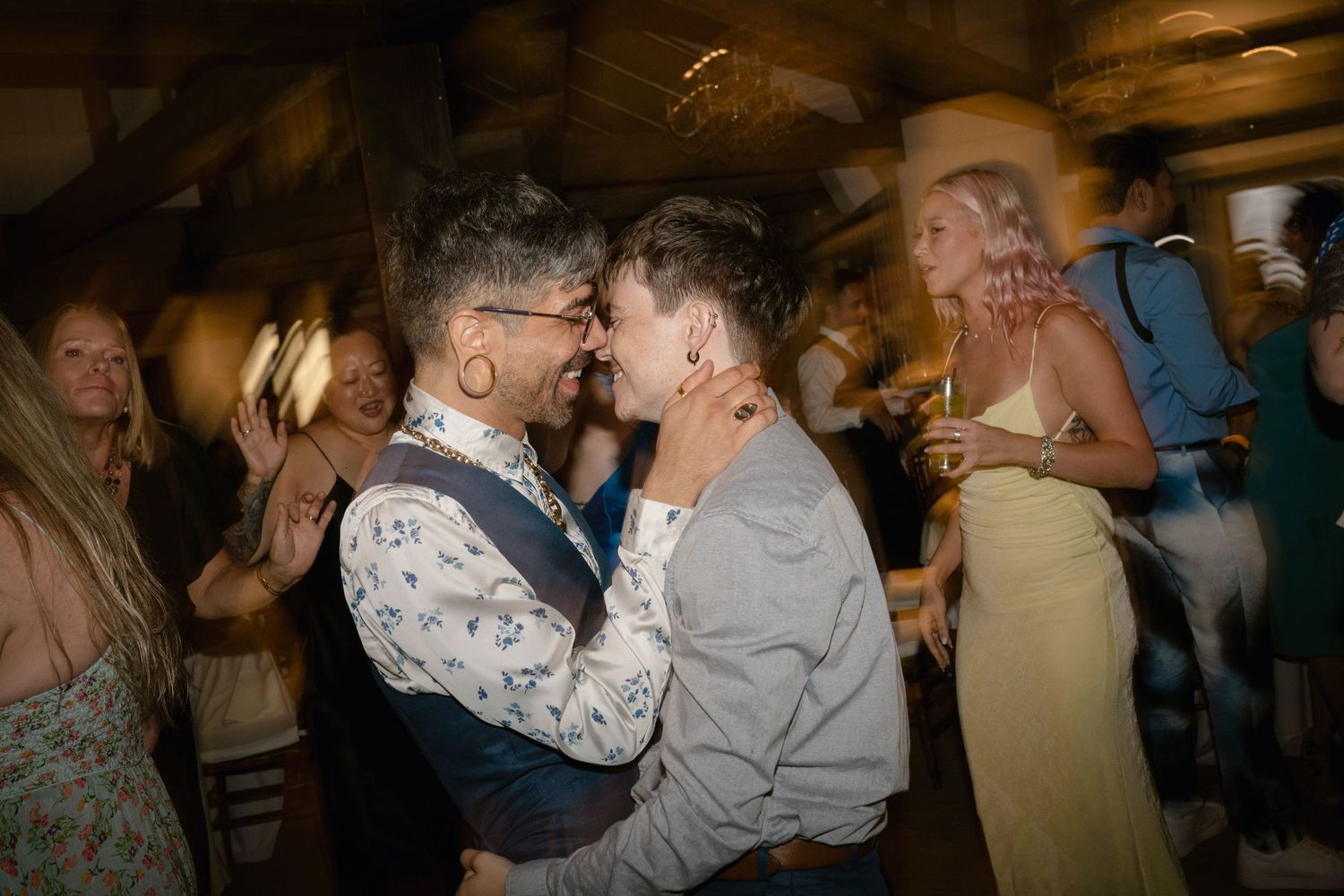Party guests embrace while dancing at a dimly lit indoor celebration.